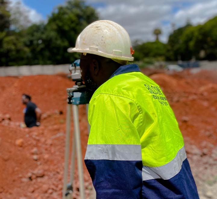 A construction worker wearing a white hard hat and high-visibility jacket looks through a surveying instrument on a tripod at a construction site with red soil.
