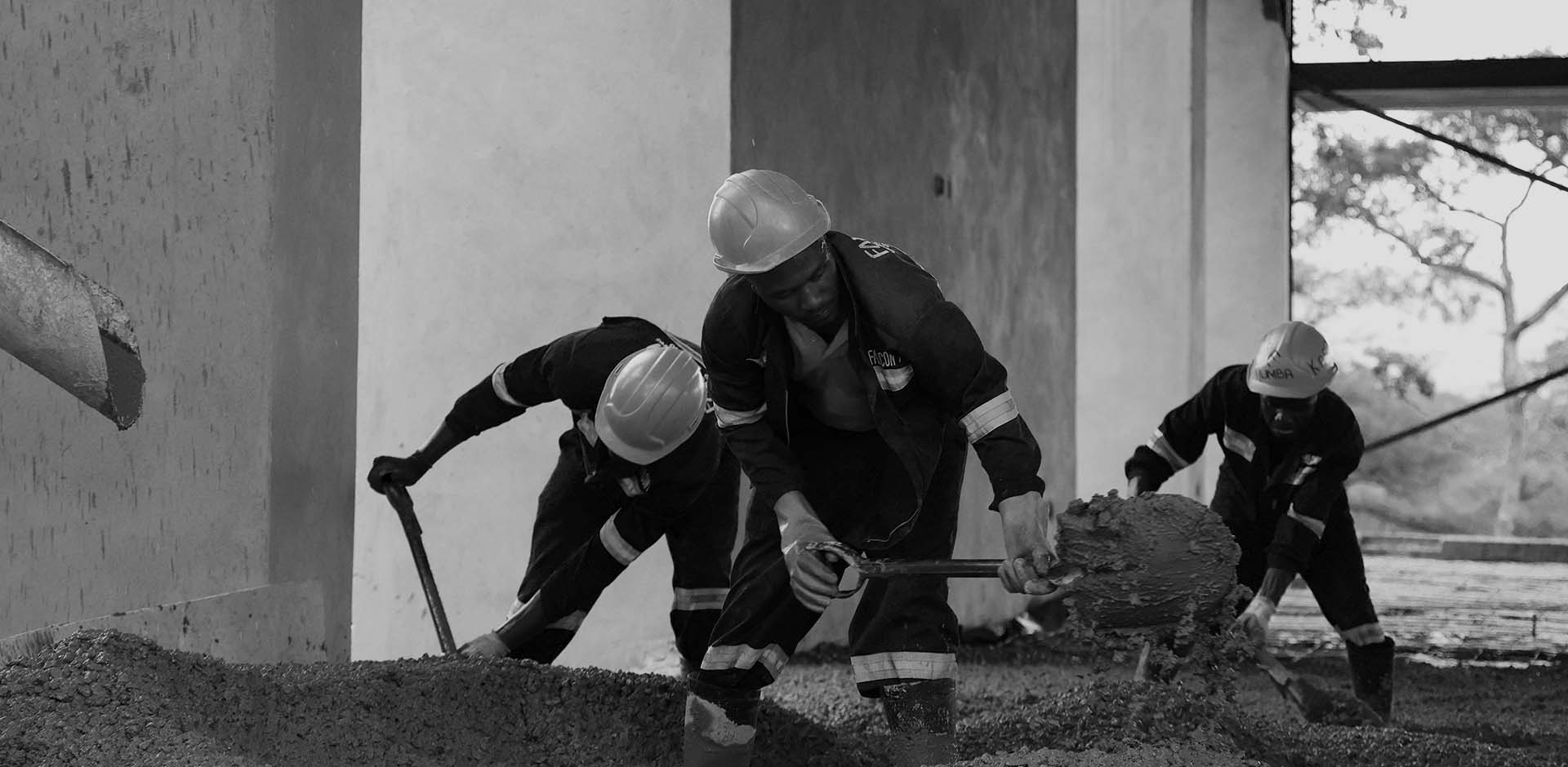 Three construction workers wearing hard hats and dark workwear are shoveling concrete mixture indoors in a partially built structure with an open window showing trees outside.