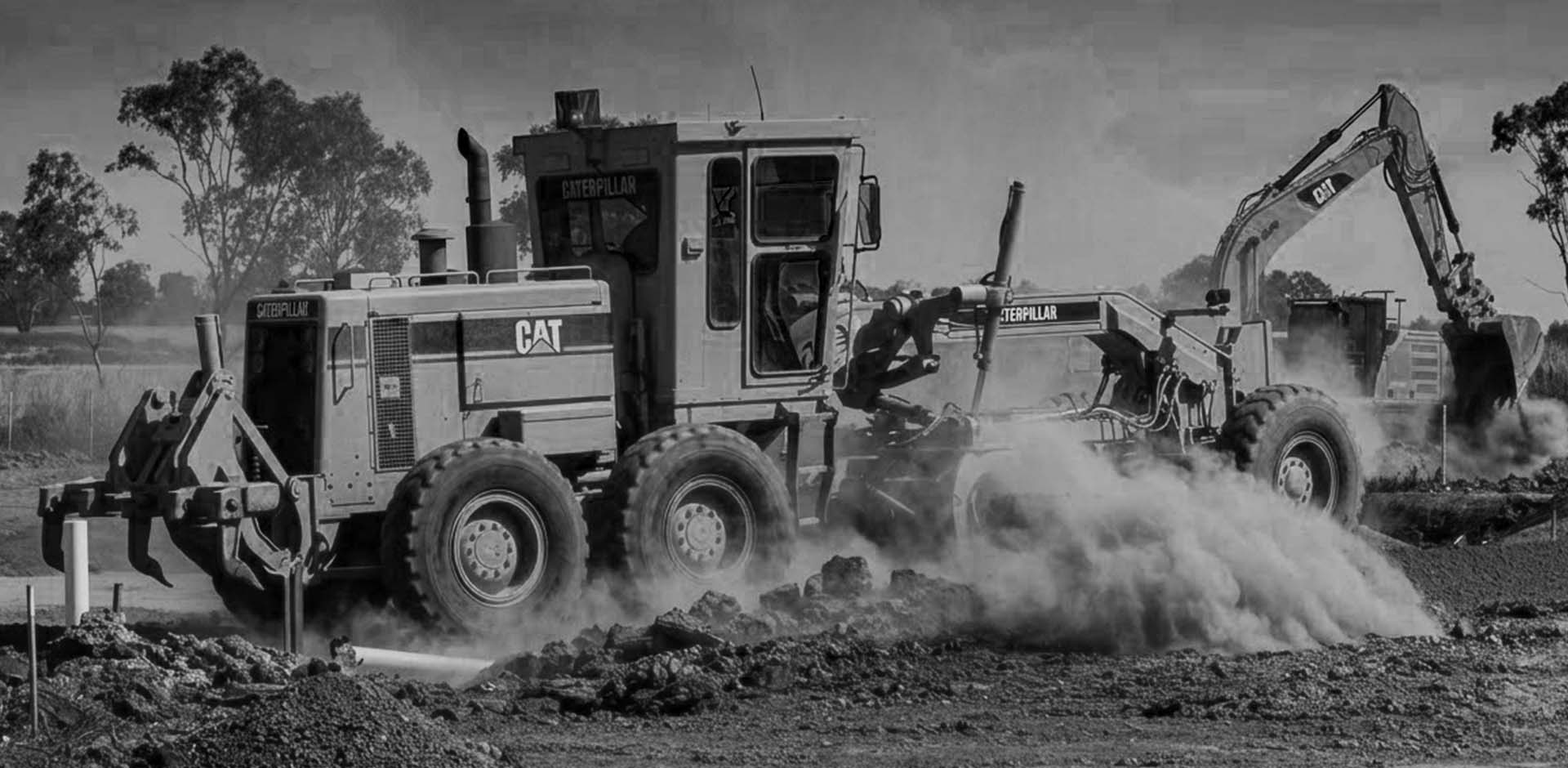 A Caterpillar motor grader on a dusty construction site leveling soil with trees and an excavator in the background.