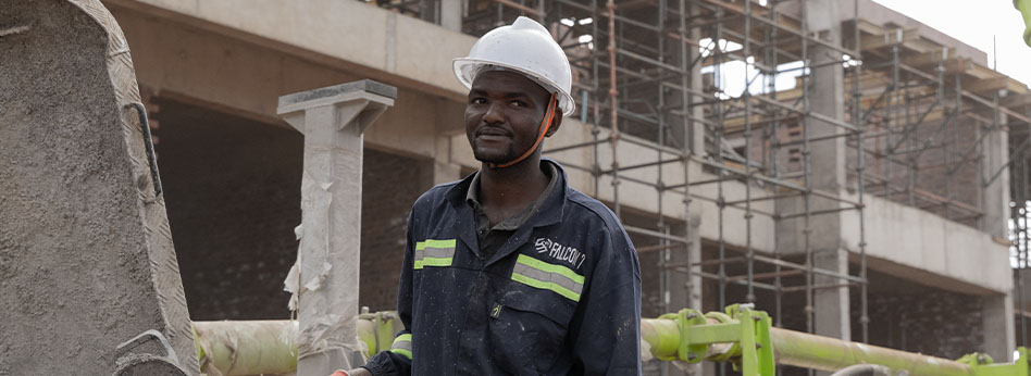 Construction worker wearing a white hard hat and dark jacket with reflective stripes stands in front of a building under construction with scaffolding.