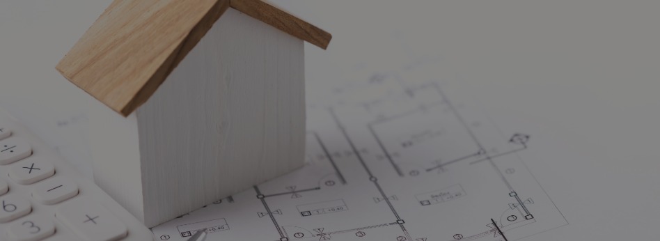 Small wooden model of a house with a slanted roof placed on a blueprint of a building plan, next to a white calculator, representing architecture or home finance concepts.