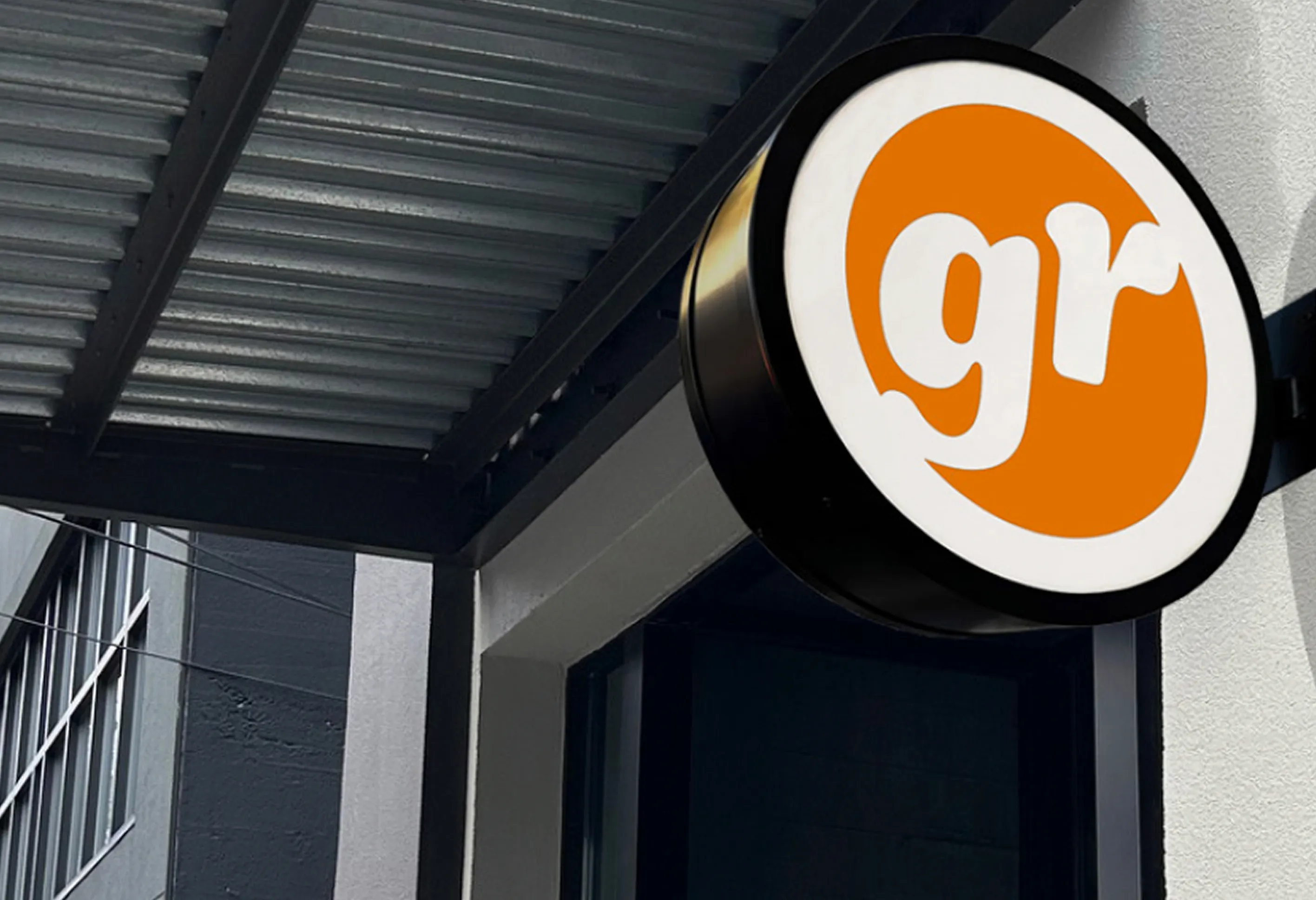 Round wall-mounted sign with a white background and orange circle containing the lowercase letters 'gr' in white, attached to the exterior of a building with a metal awning.