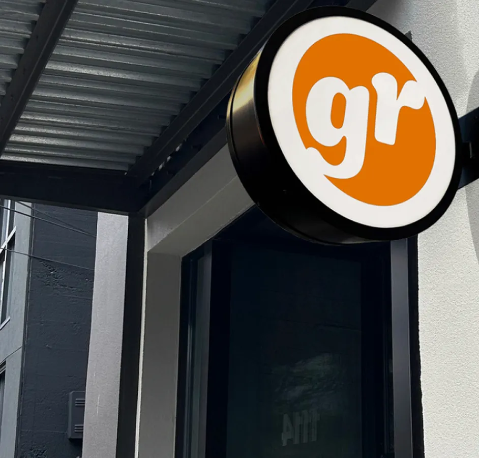 Round sign with white lowercase letters 'gr' on an orange circle, mounted on the corner of a building under a metal overhang.