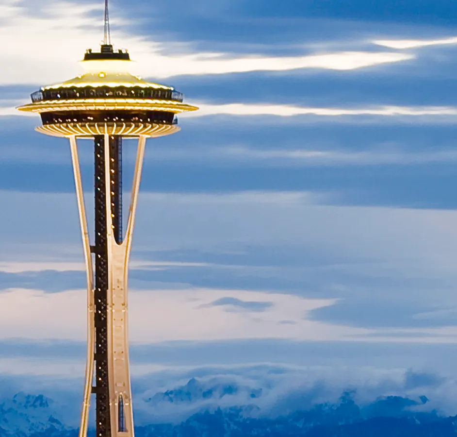 The illuminated Space Needle tower in Seattle against a cloudy blue sky with a mountain range faintly visible at the bottom.