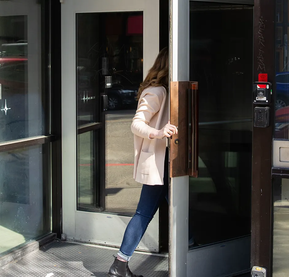 Person wearing a beige jacket and dark jeans opening a glass door with a wooden handle, stepping outside onto a sidewalk.
