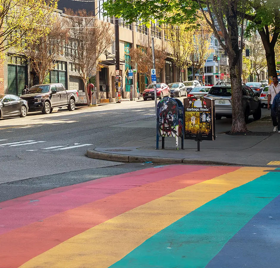 City street scene with a crosswalk painted in rainbow colors, symbolizing LGBTQ pride, alongside parked cars, trees, and two mailboxes near the sidewalk.