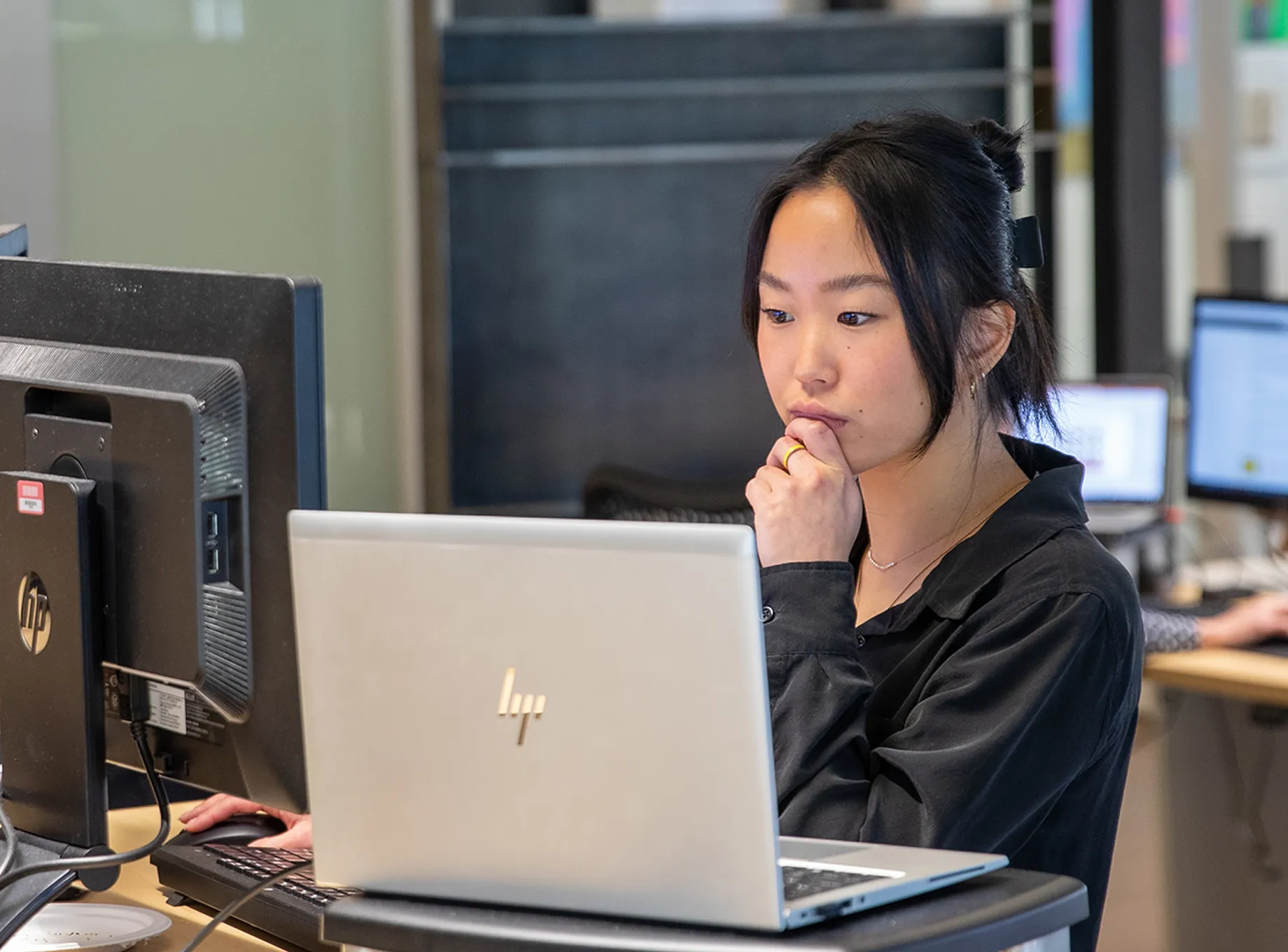 Young woman with dark hair tied back, wearing a black shirt, sitting at a desk working on a silver HP laptop with a larger monitor beside it in an office environment.