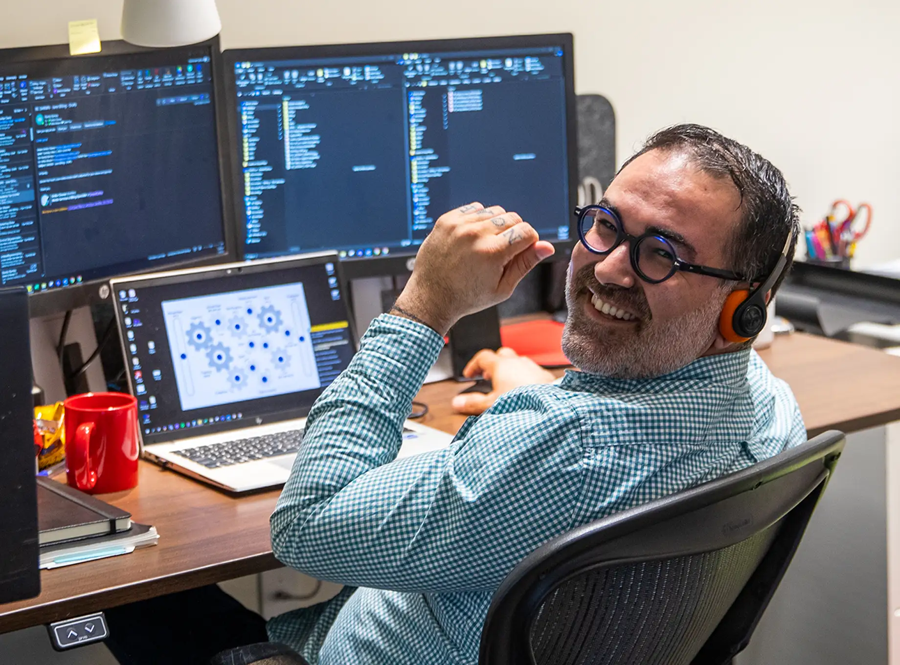 A man wearing round glasses and headphones smiles while sitting at a desk with multiple computer screens displaying code and diagrams in a workspace.