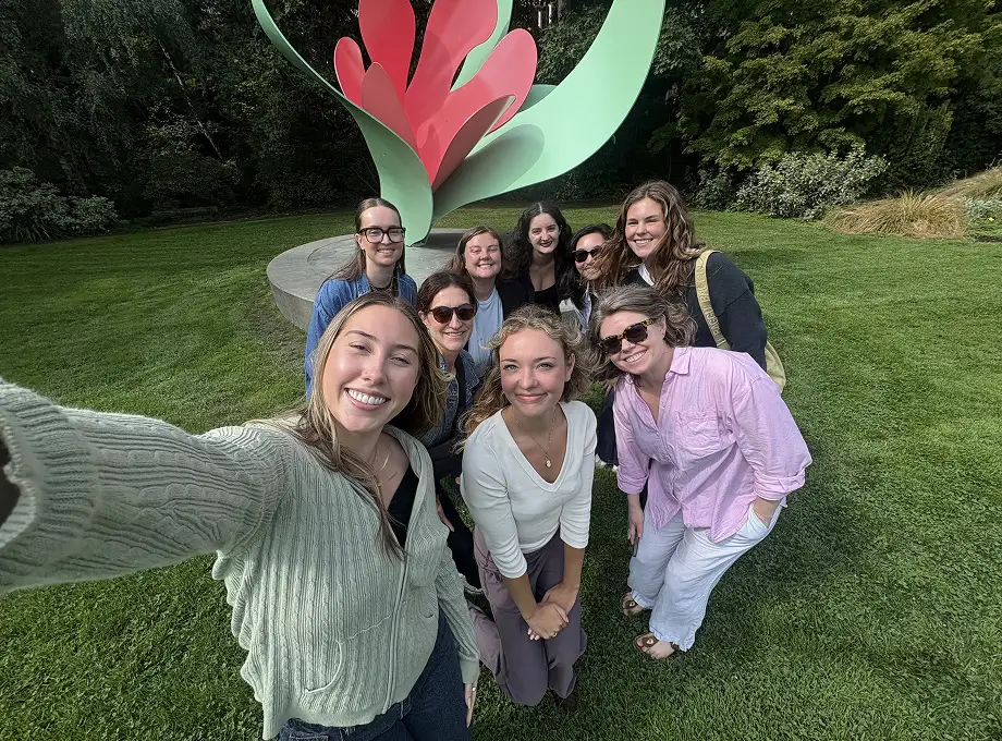 Group of nine smiling women taking a selfie outdoors on green grass with a large abstract red and green sculpture and trees in the background.