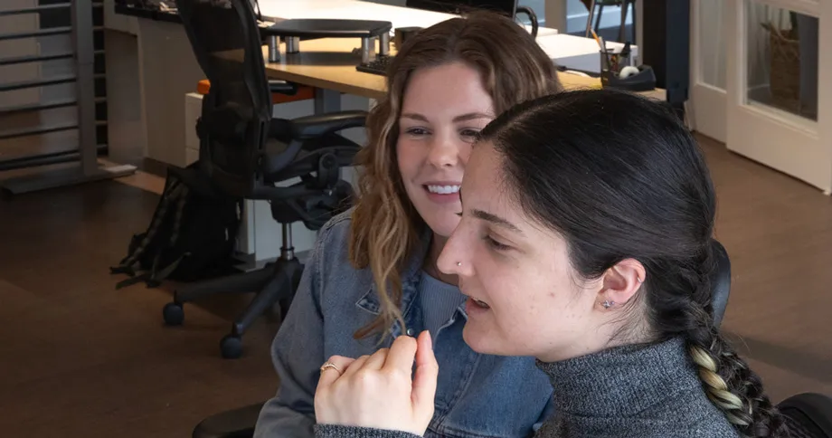 Two women sitting closely together in an office setting, one with braided dark hair wearing a gray turtleneck sweater and the other with wavy light brown hair wearing a denim jacket, both appearing engaged and smiling.