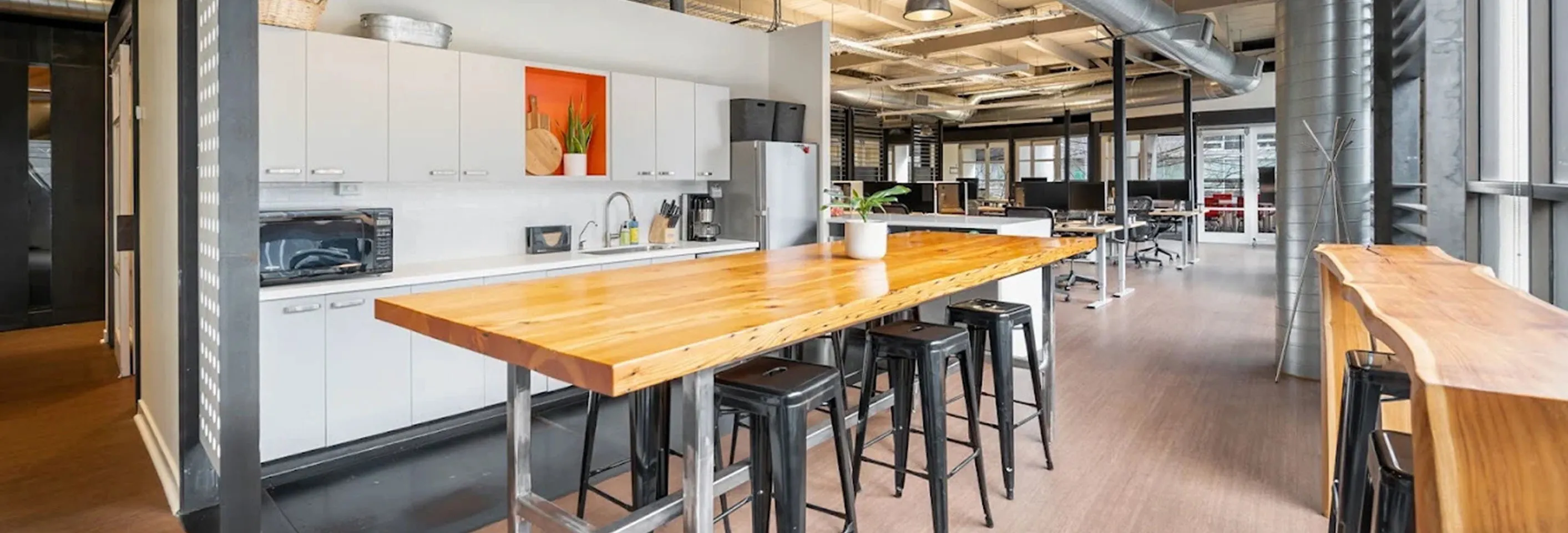 Modern office kitchen with a long wooden island table, black stools, white cabinets, a microwave, coffee maker, and stainless steel refrigerator, with an open office workspace in the background.