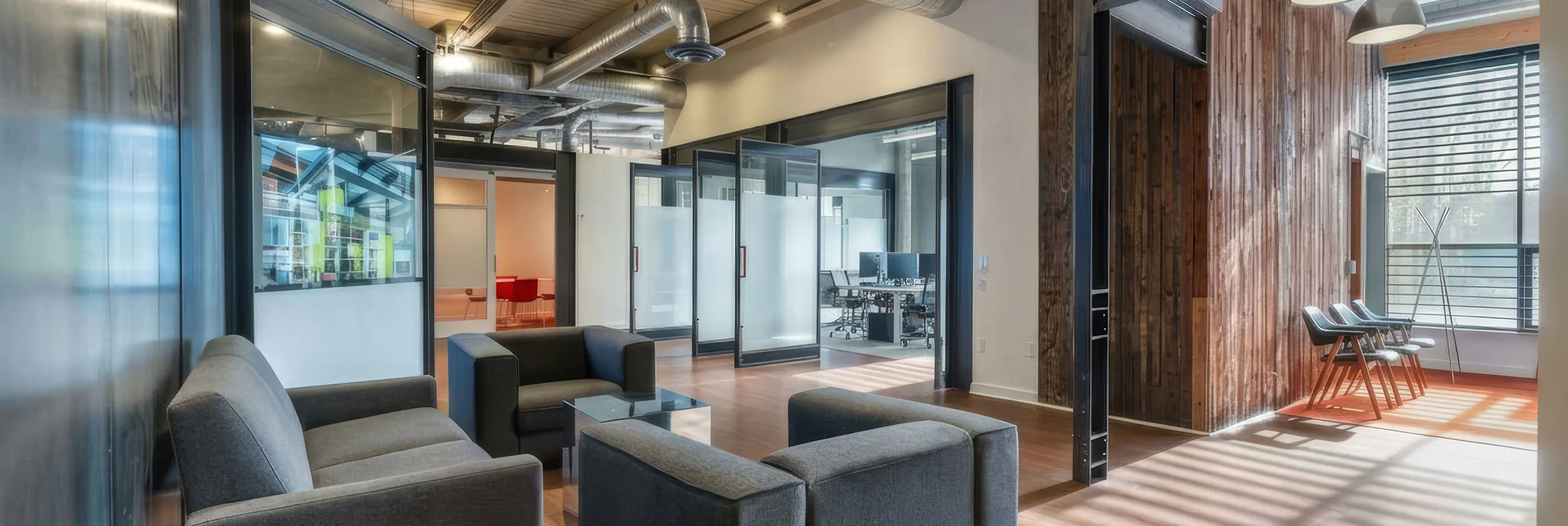 Modern office lounge area with grey upholstered armchairs arranged around a glass coffee table, adjacent to glass-walled conference rooms and workspaces, featuring wood-paneled walls and large windows letting in natural light.