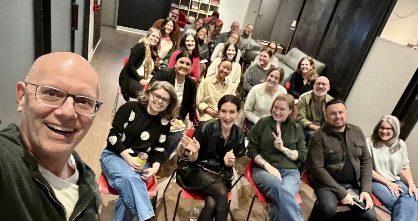 A group of smiling adults seated in rows of red chairs facing the camera, with a man in glasses taking a selfie in the foreground inside a room with dark walls and shelves in the background.