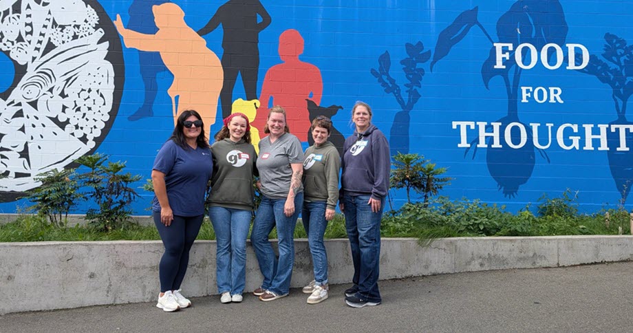 Five women standing side by side smiling in front of a vibrant blue mural with colorful silhouettes of people, plants, fish, and the words 'FOOD FOR THOUGHT'.