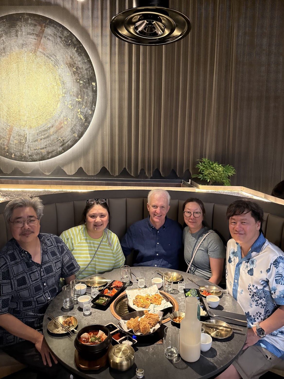 Five friends around a Korean grill table at Soom in London, mid-meal, with banchan, fried chicken, stew and makgeolli
