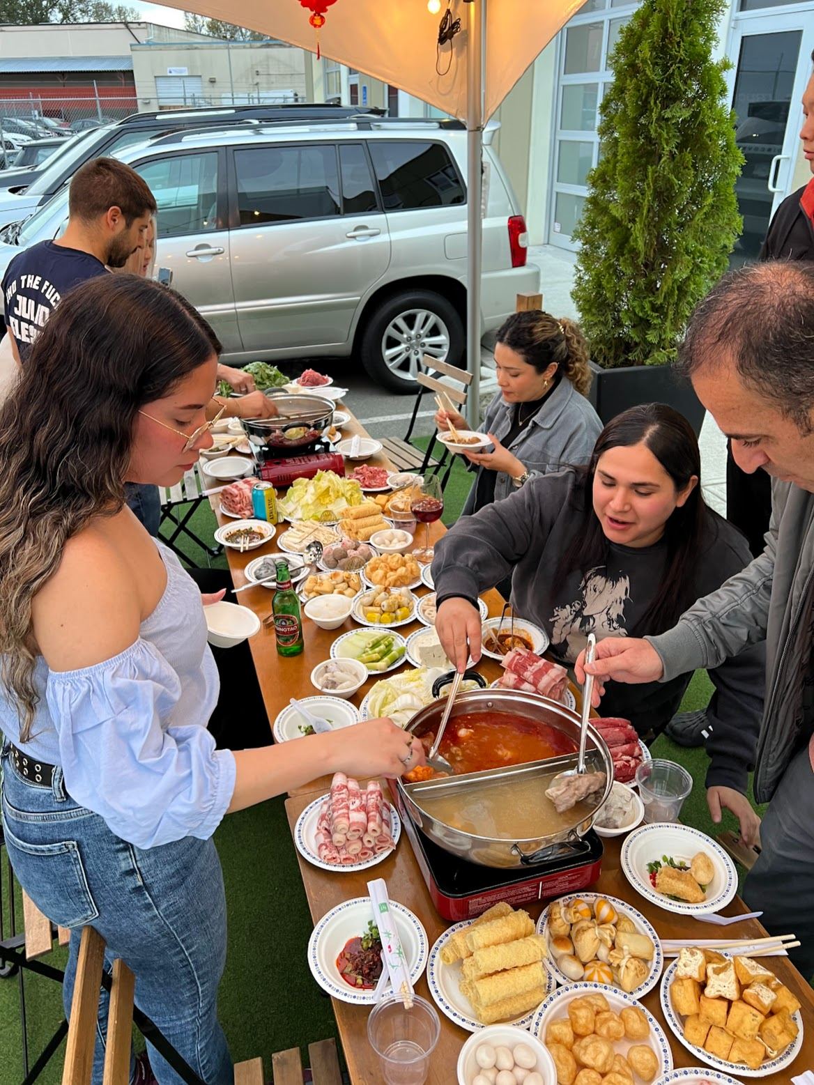 Friends and family around a long outdoor table sharing hot pot on Lunar New Year, tongs reaching into a split broth surrounded by plates of rolled meat, tofu and vegetables