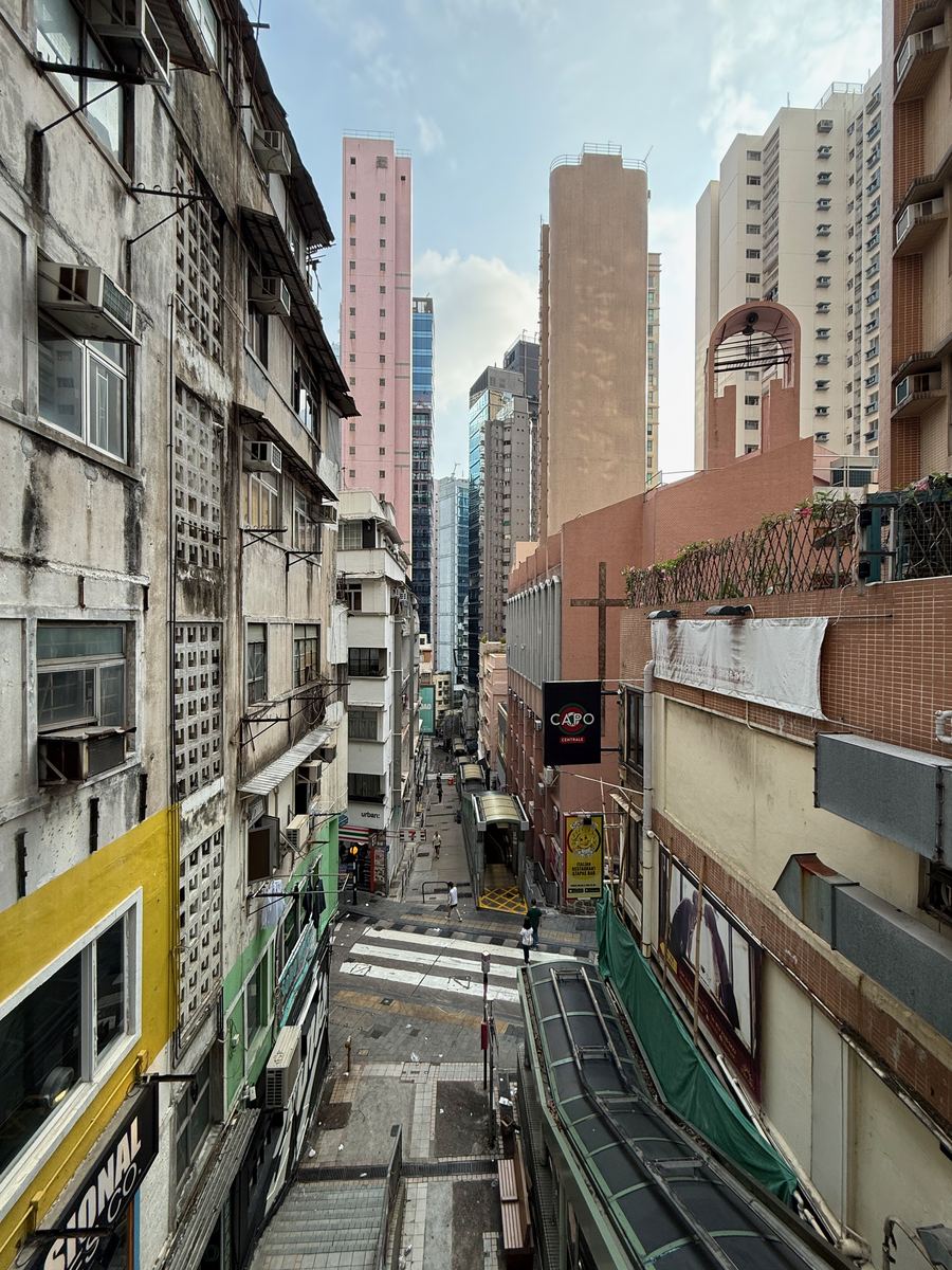 A narrow Hong Kong street seen from above early in the morning, tenement blocks and shop signs on both sides and towers rising behind