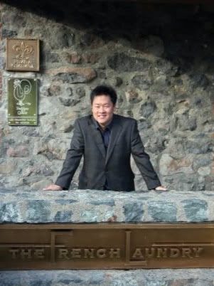 A man in a dark suit leaning on the low stone wall at the entrance to The French Laundry in Yountville, California, the restaurant's name carved into the sign below him