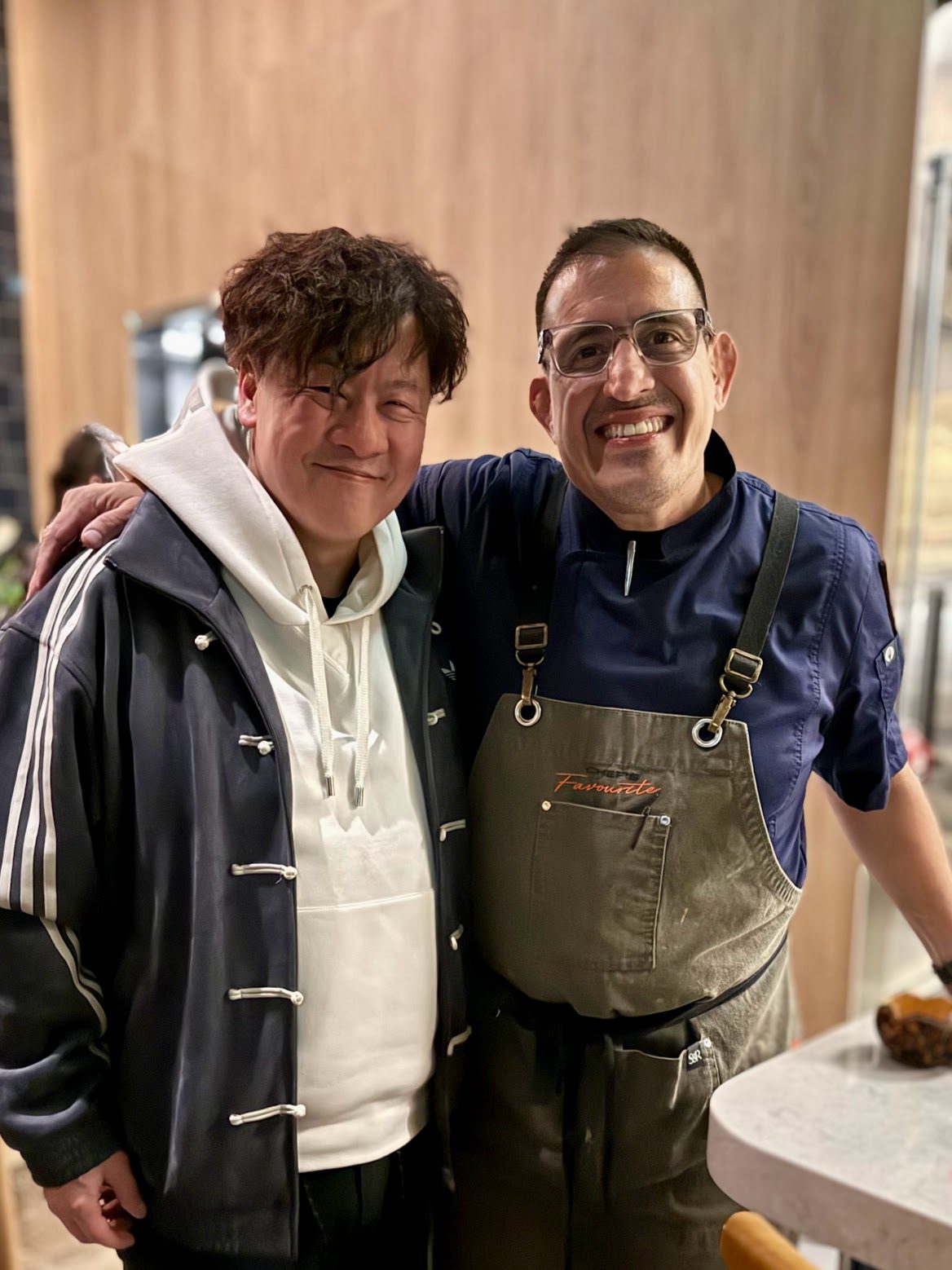 A chef in a navy shirt and canvas apron standing arm in arm with a friend in a restaurant dining room, both smiling at the camera