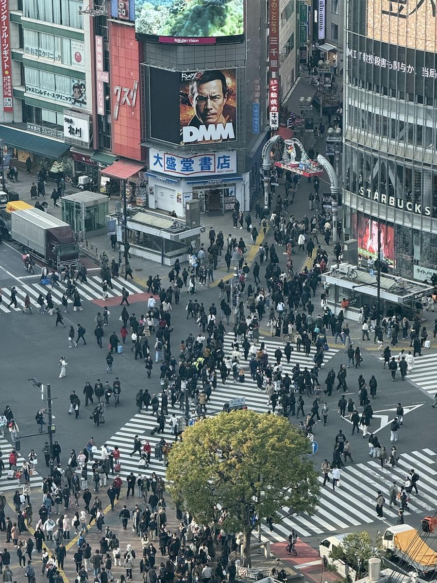 Shibuya Crossing photographed from high inside the station complex, crowds streaming across the diagonal crosswalks between illuminated billboards and department stores