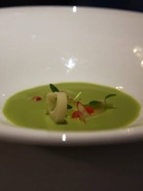 A shallow white bowl of pale green cucumber gazpacho with rings of peach palm, halved grapes, red radish and cilantro shoots