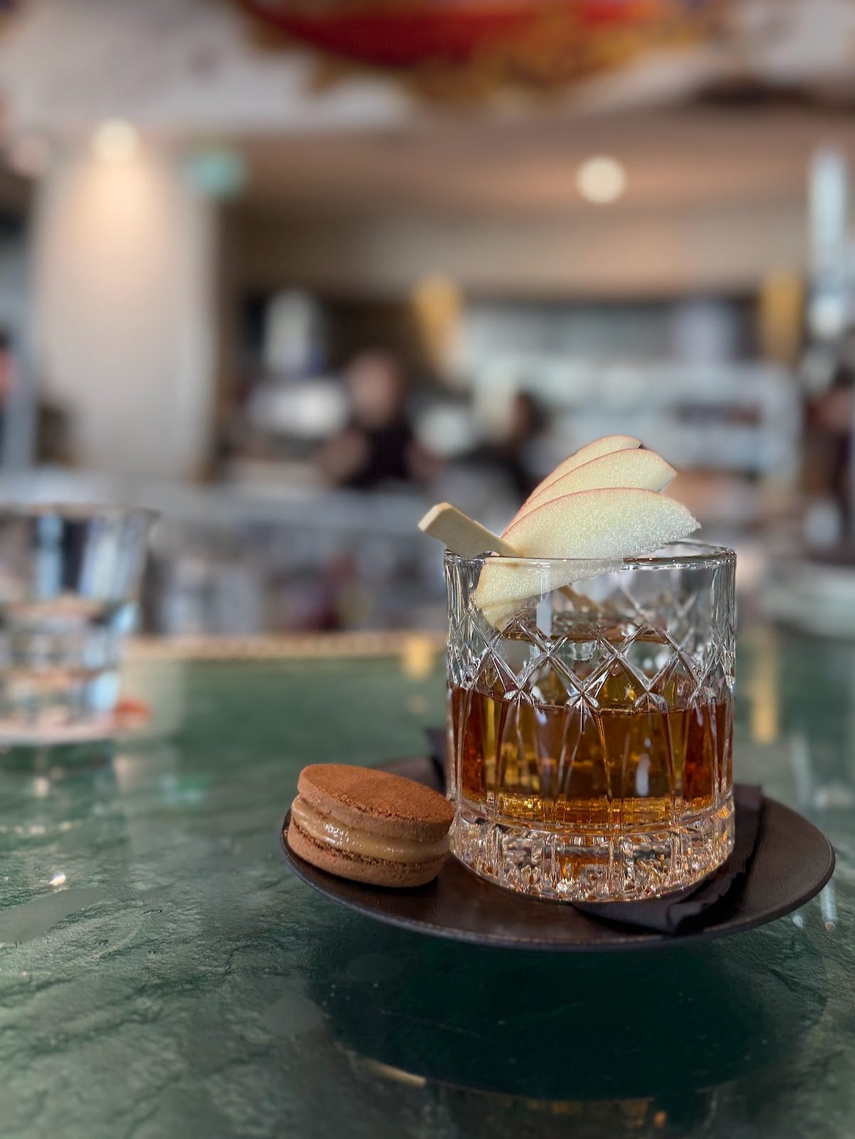 An amber cognac cocktail in a cut-crystal glass garnished with a fan of apple slices, with a chocolate macaron on a small plate beside it