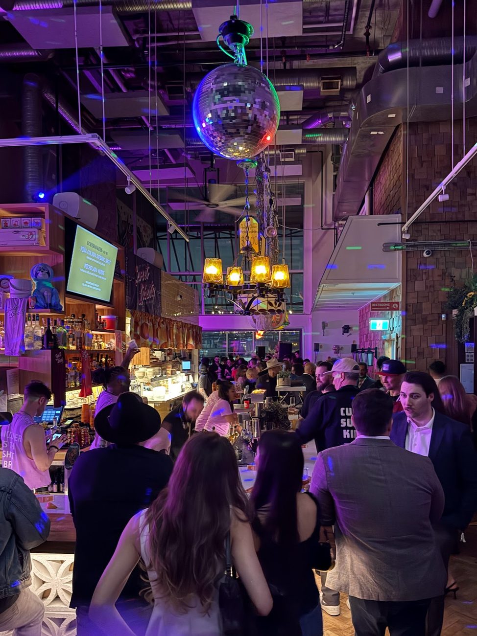 A crowded late-night bar under a disco ball and brass chandelier, a cocktail menu on a screen behind the bar