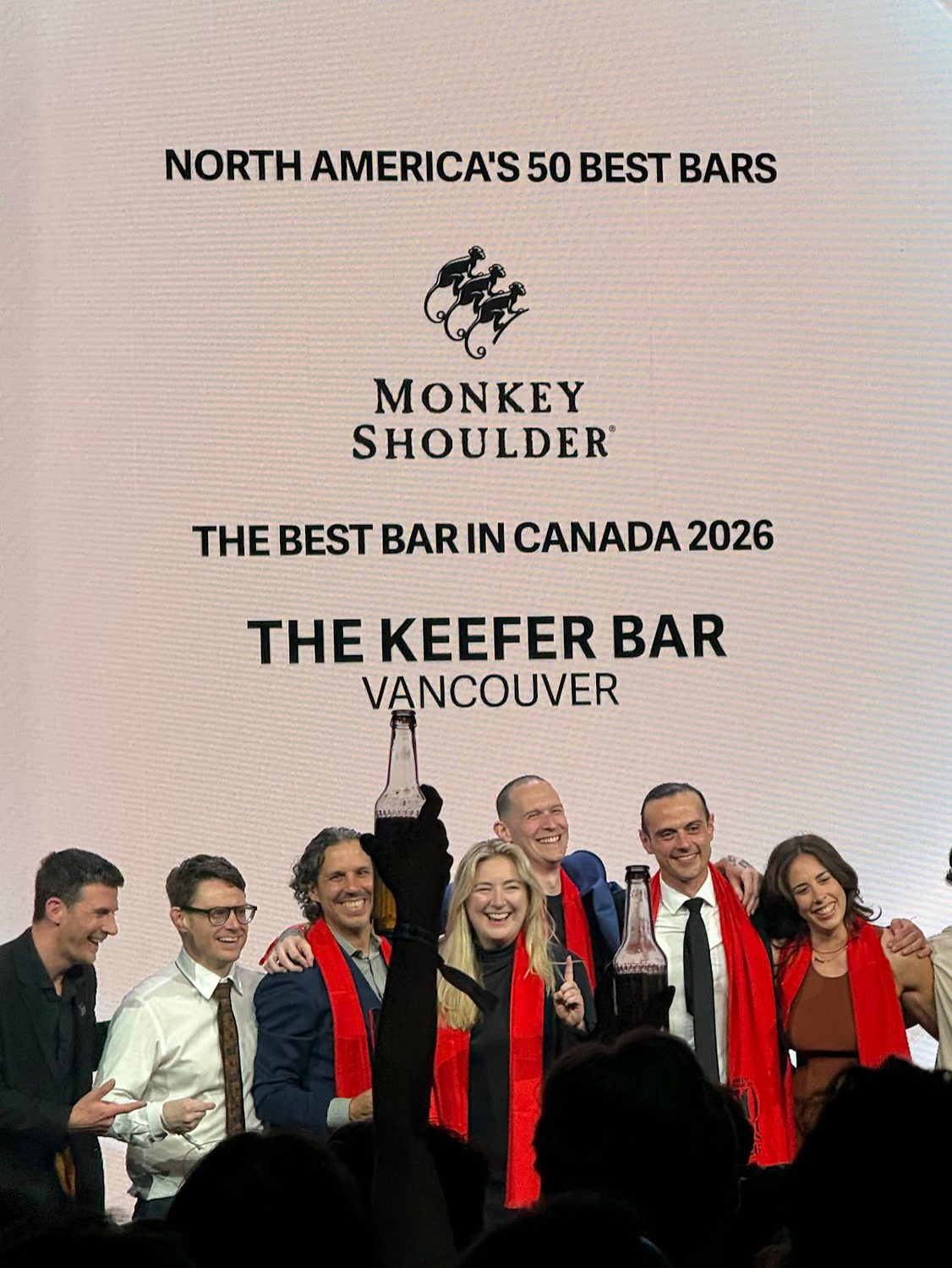 The Keefer Bar team celebrating on stage in red scarves beneath a screen reading Monkey Shoulder The Best Bar in Canada 2026