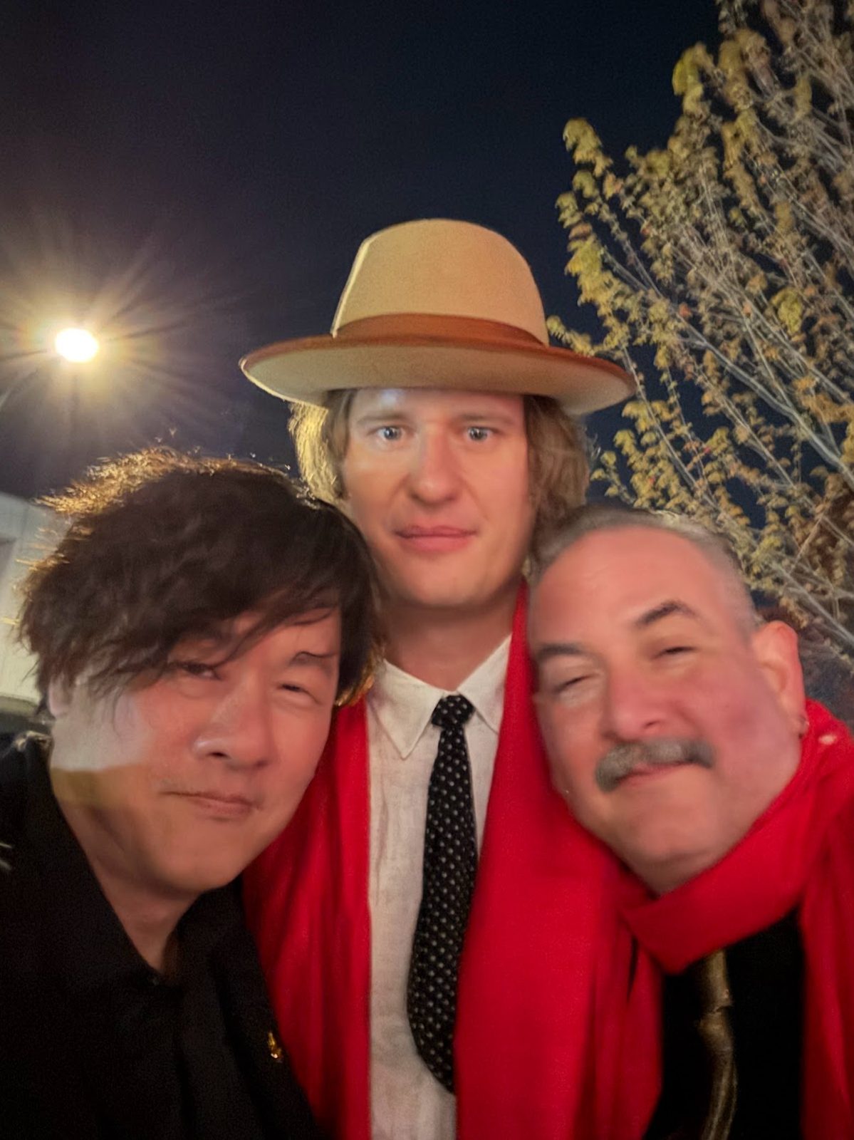 Three men outdoors at night under a streetlight, two wearing red 50 Best scarves and one in a tan fedora
