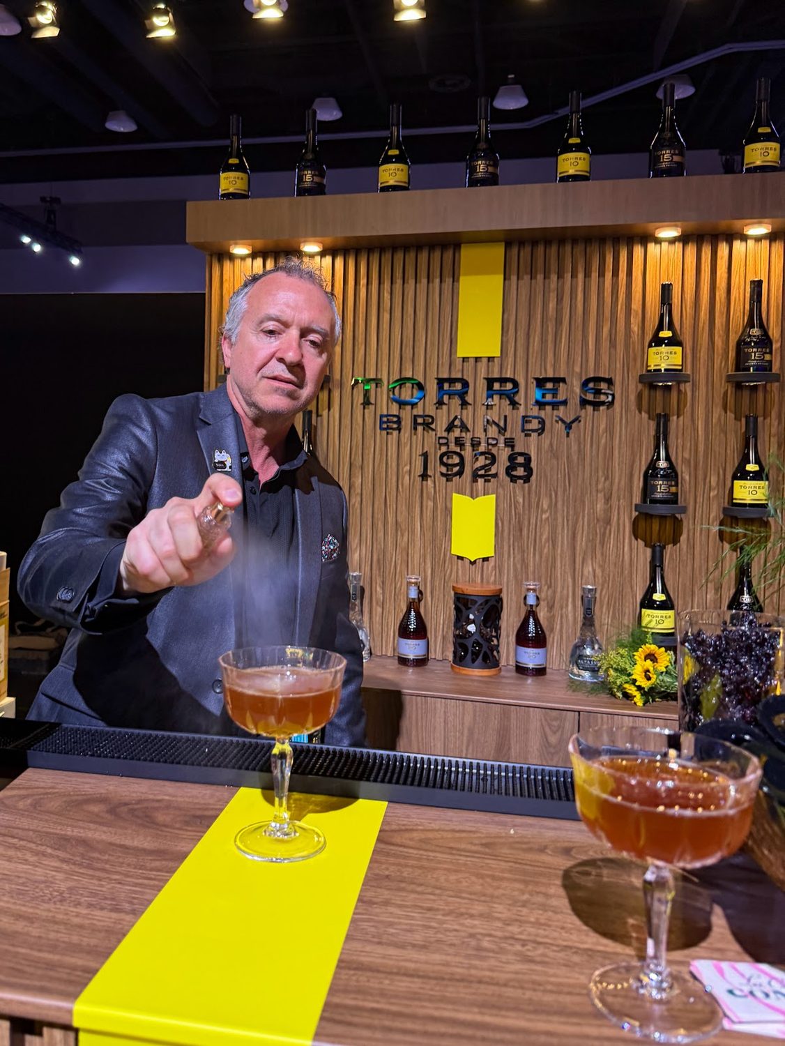 A bartender misting the surface of a coupe at the Torres Brandy bar, bottles ranged on the wooden back bar behind him
