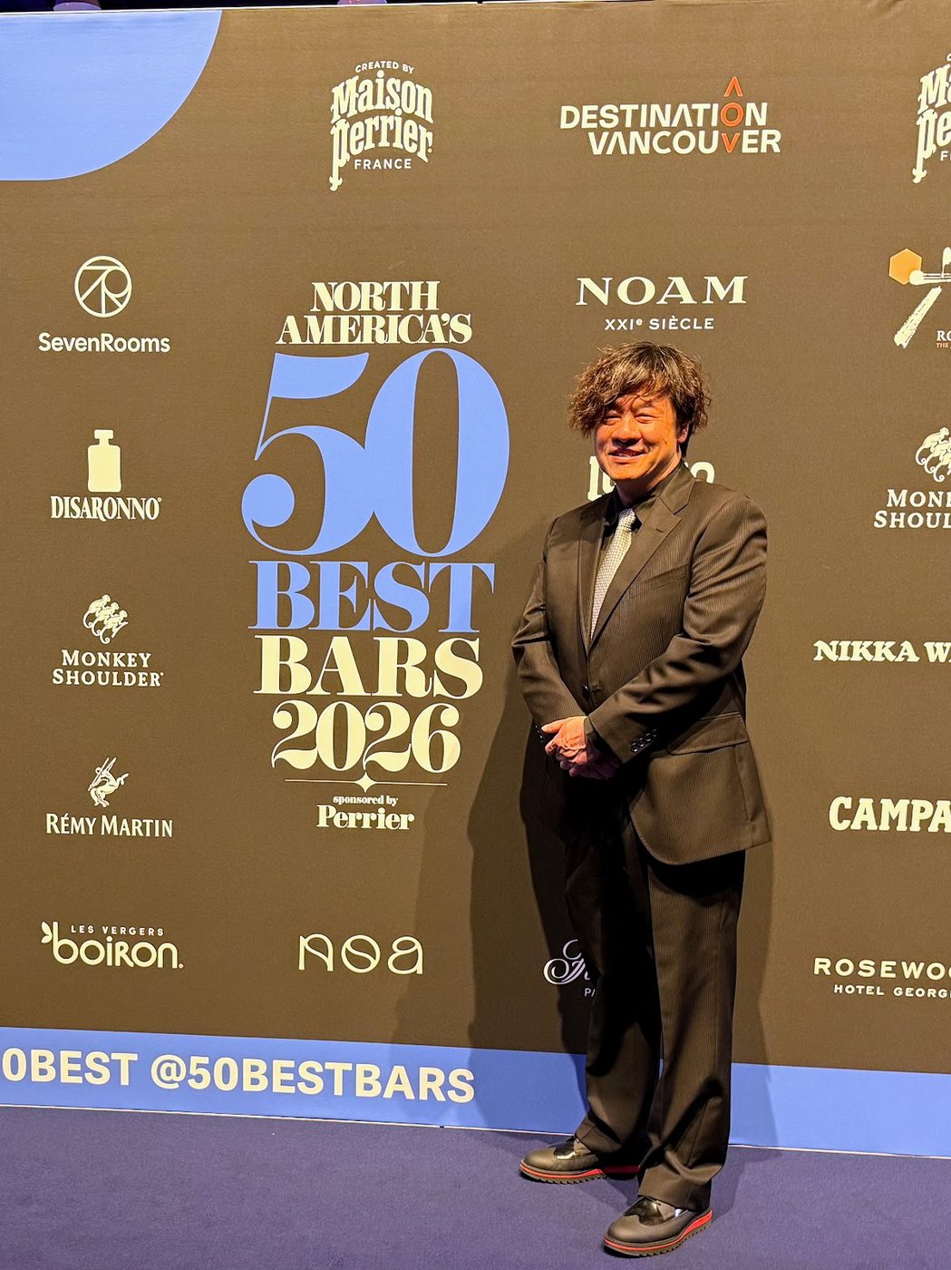 A guest in a brown pinstripe suit standing in front of the North America's 50 Best Bars 2026 sponsor backdrop