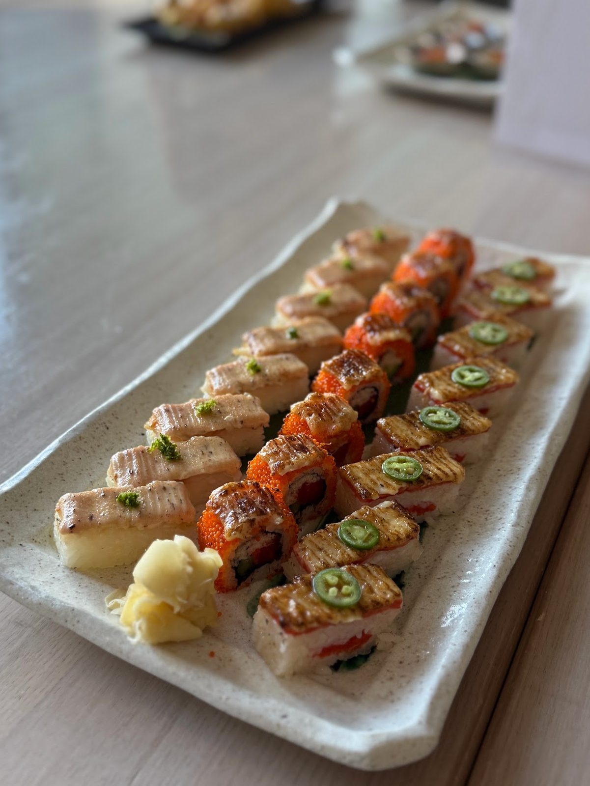 A long white ceramic platter of flame-seared pressed sushi in three rows, topped with jalapeño slices and orange tobiko, with pickled ginger alongside