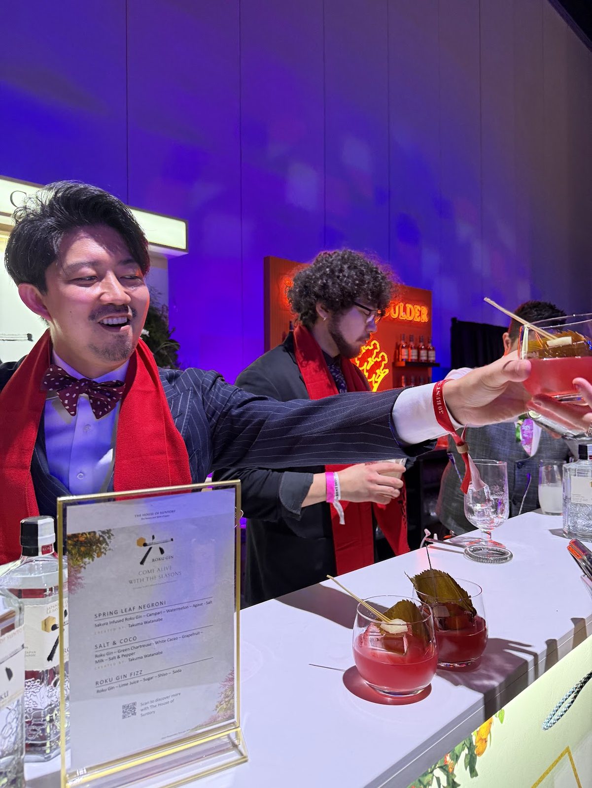 A bartender in a bow tie and red scarf passing a garnished cocktail across the Roku Gin bar, with the printed drinks menu in a brass frame in the foreground