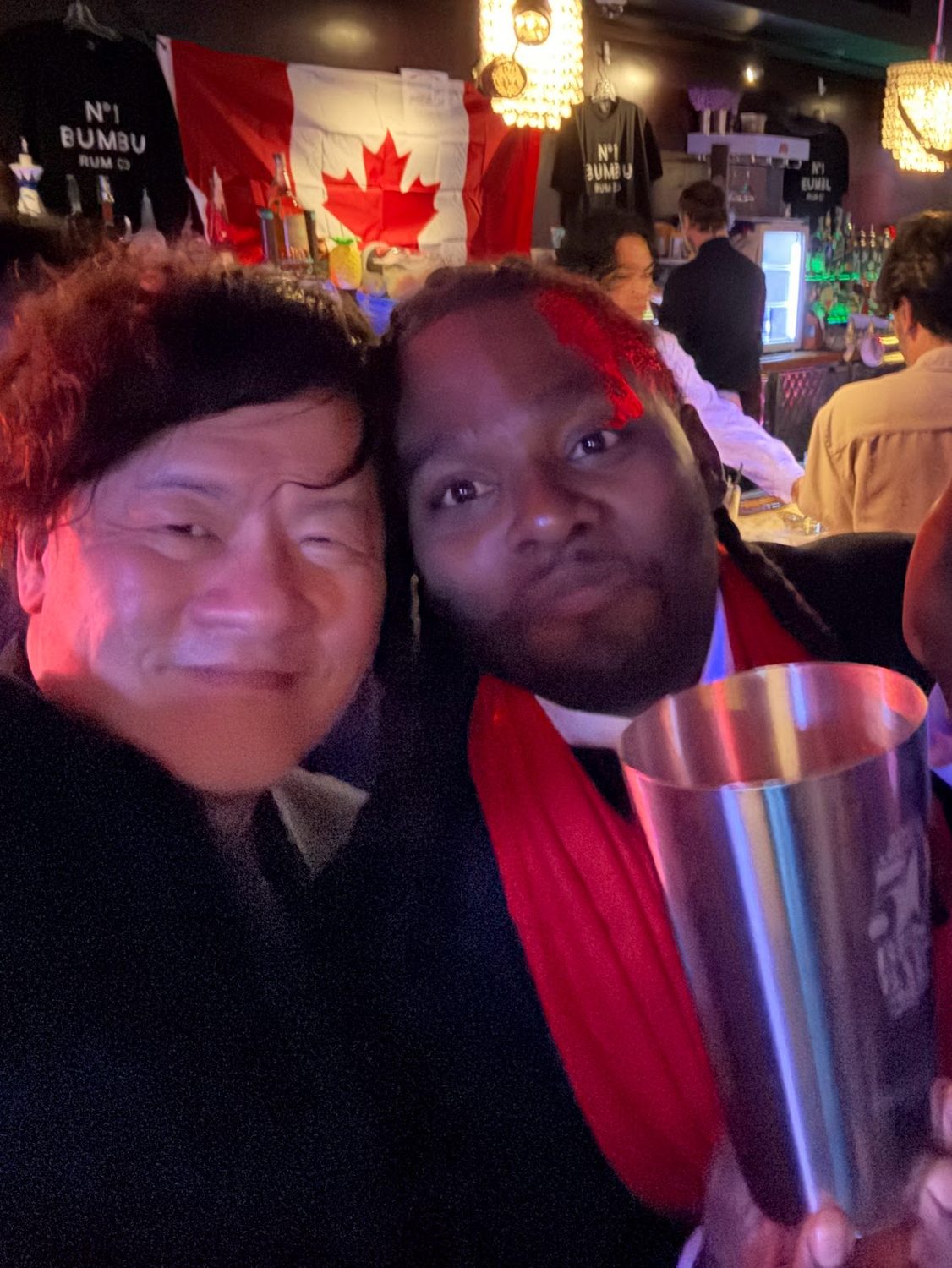 A bartender in a red scarf holding a shaker tin at a small bar hung with a Canadian flag and rum T-shirts