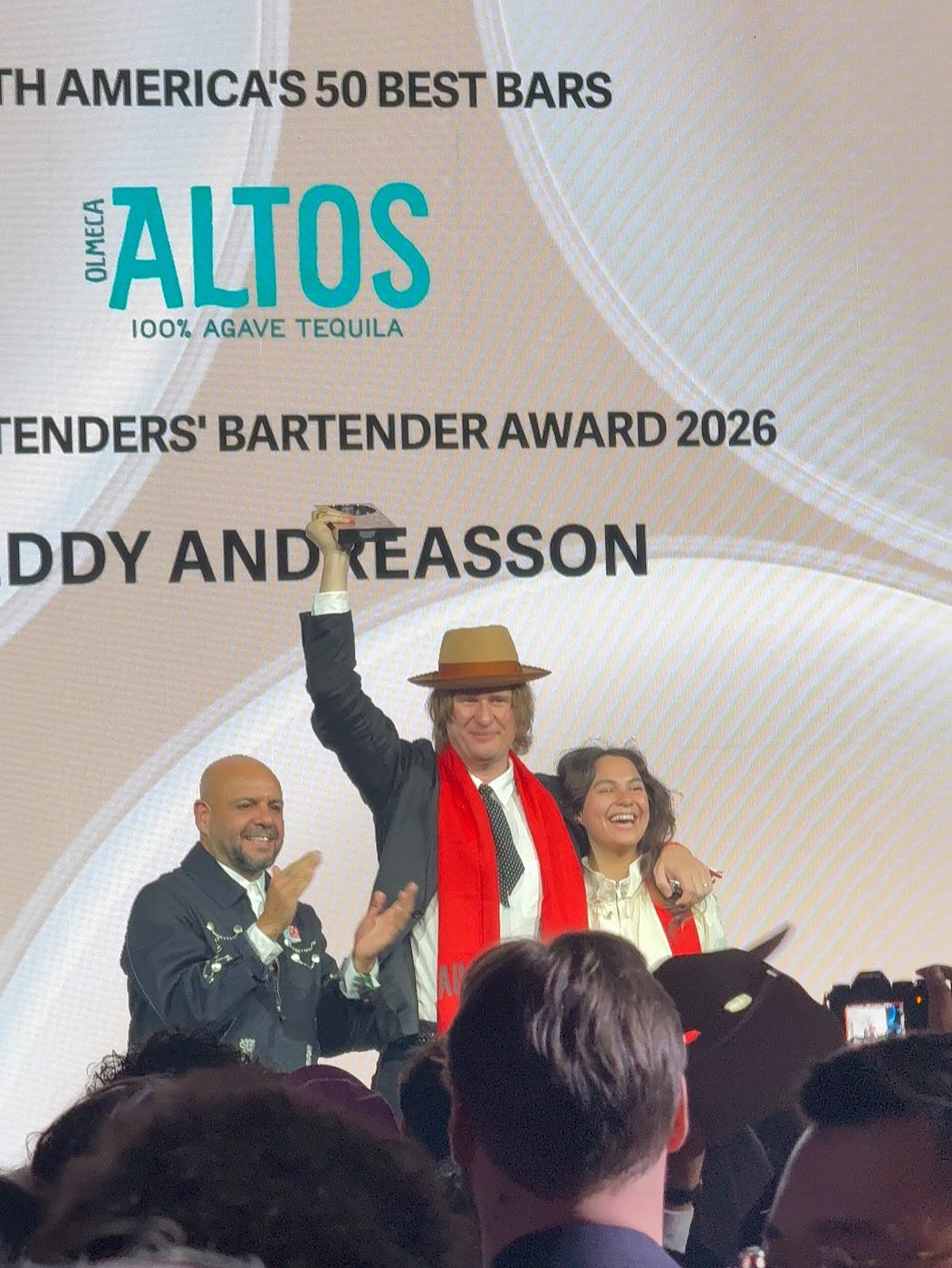A bartender in a tan fedora and red scarf holding his trophy aloft on stage, beneath a screen reading Olmeca Altos Bartenders' Bartender Award 2026