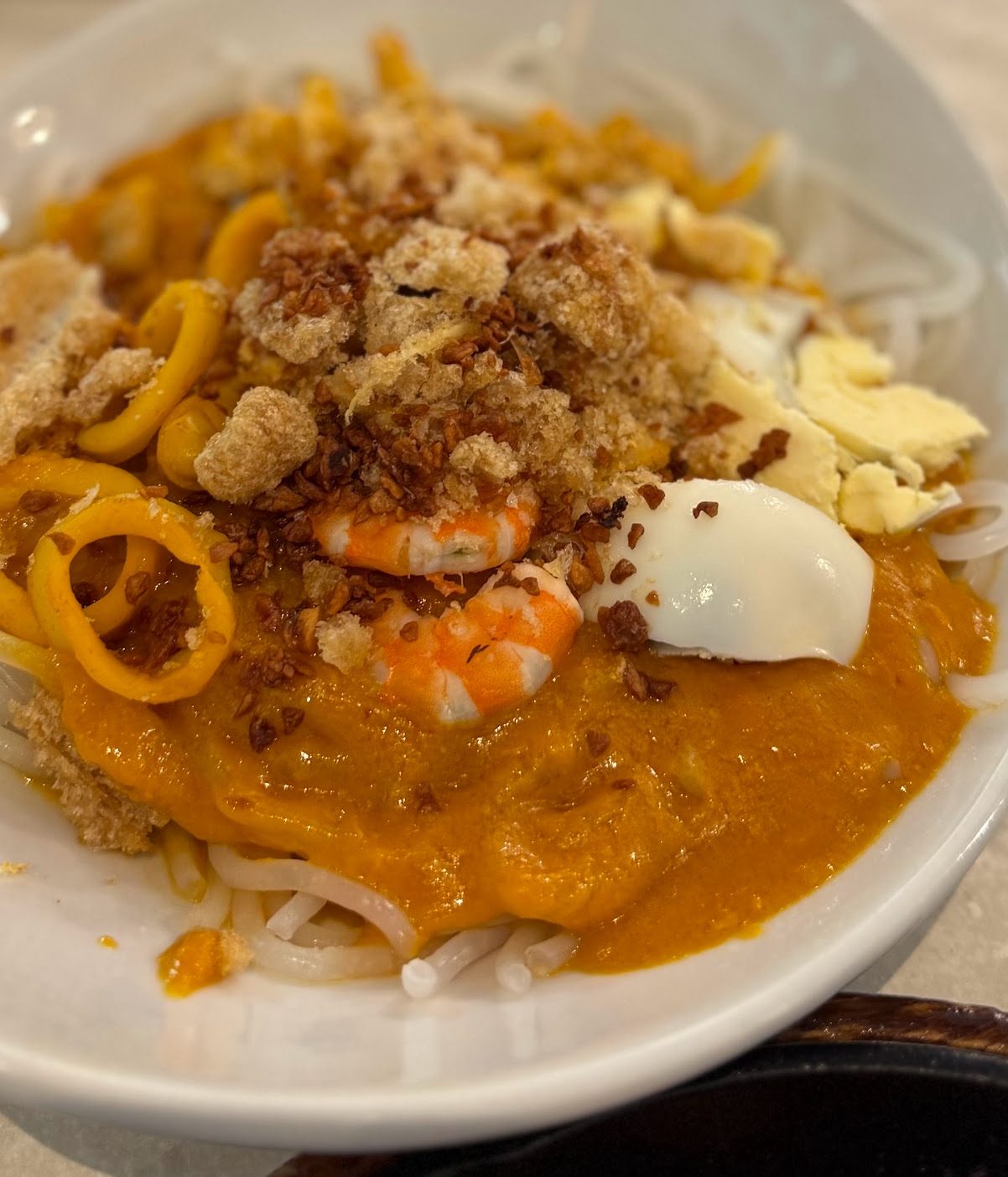 A plate of Pancit Malabon with thick rice noodles and squid rings visible beneath orange crab-fat sauce, shrimp, a boiled egg, crushed chicharon and toasted garlic