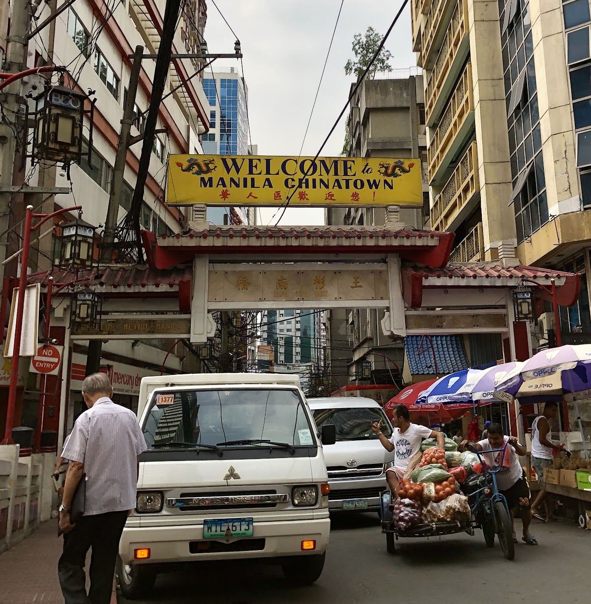 The Ongpin South Bridge arch marking the entrance to Binondo, the Chinatown of Manila, beneath a yellow "Welcome to Manila Chinatown" sign — overhead wires, vans in the narrow street and a sidecar piled with onions and vegetables, with glass office towers rising behind