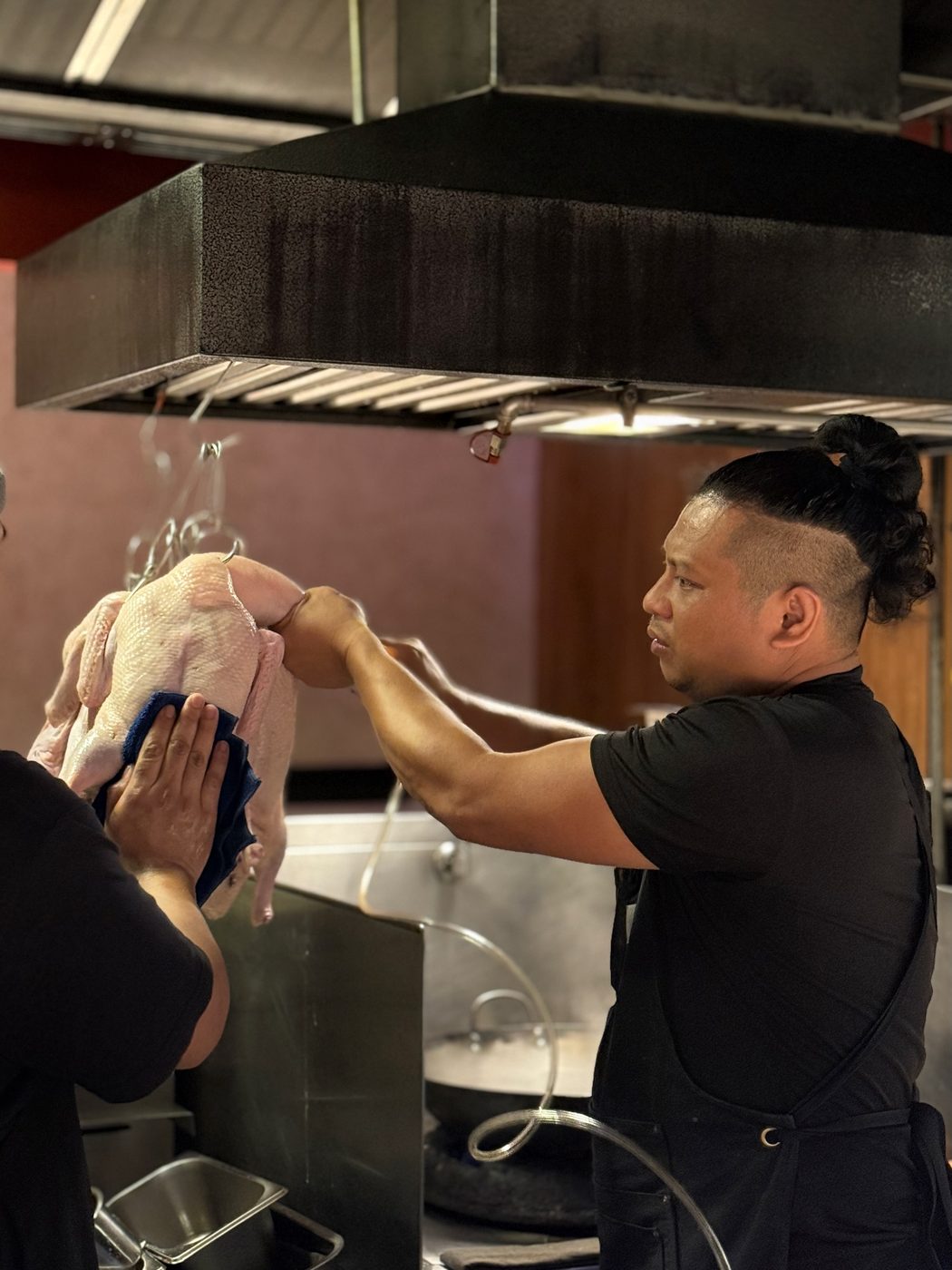 A chef in the open kitchen working compressed air under the skin of a whole raw duck, a second pair of hands steadying it, empty hanging hooks above