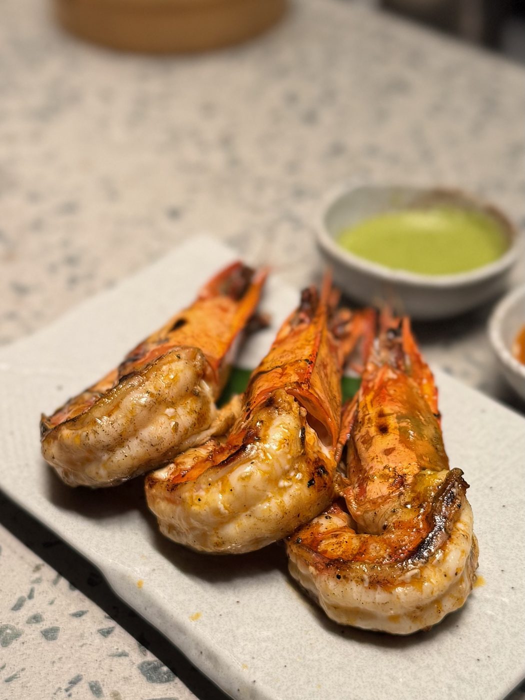 Split head-on king prawns, charred from the coals, on a ceramic plate beside a bowl of bright green sauce