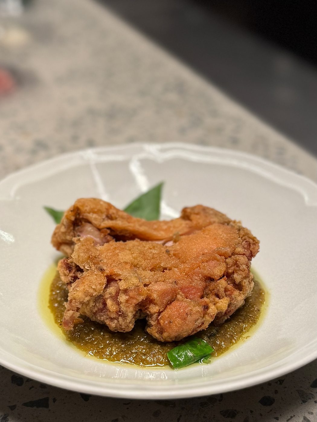 A craggy, golden brown piece of fried chicken sitting on a pool of dry green curry sauce with curry leaves