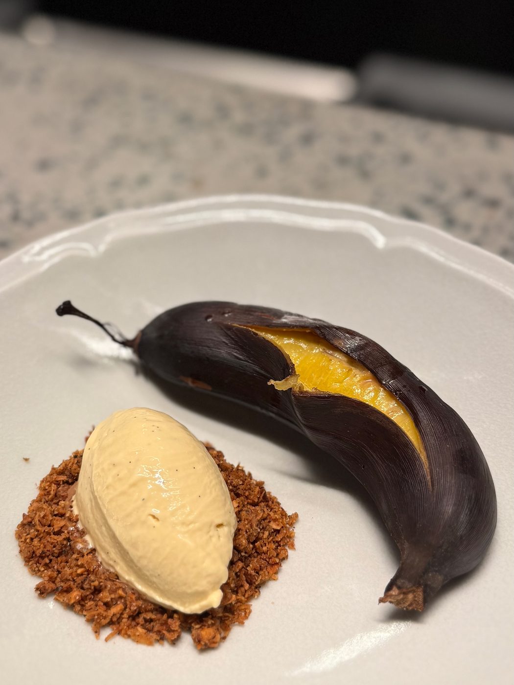 A whole banana roasted in its blackened skin and split open, served with a scoop of vanilla ice cream on toasted coconut