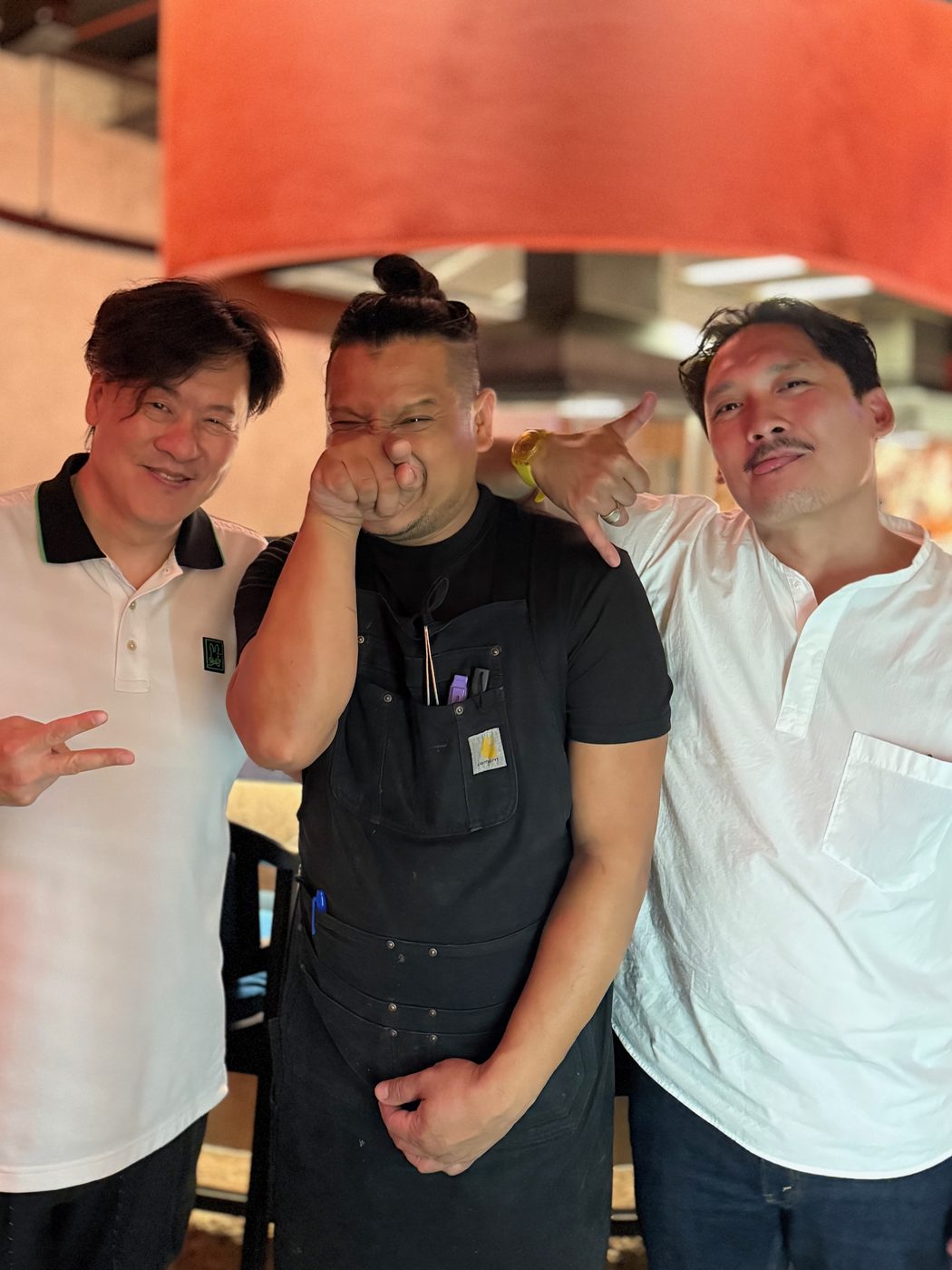 Three men standing together under an orange pendant lamp at a restaurant, one of them in a black kitchen apron
