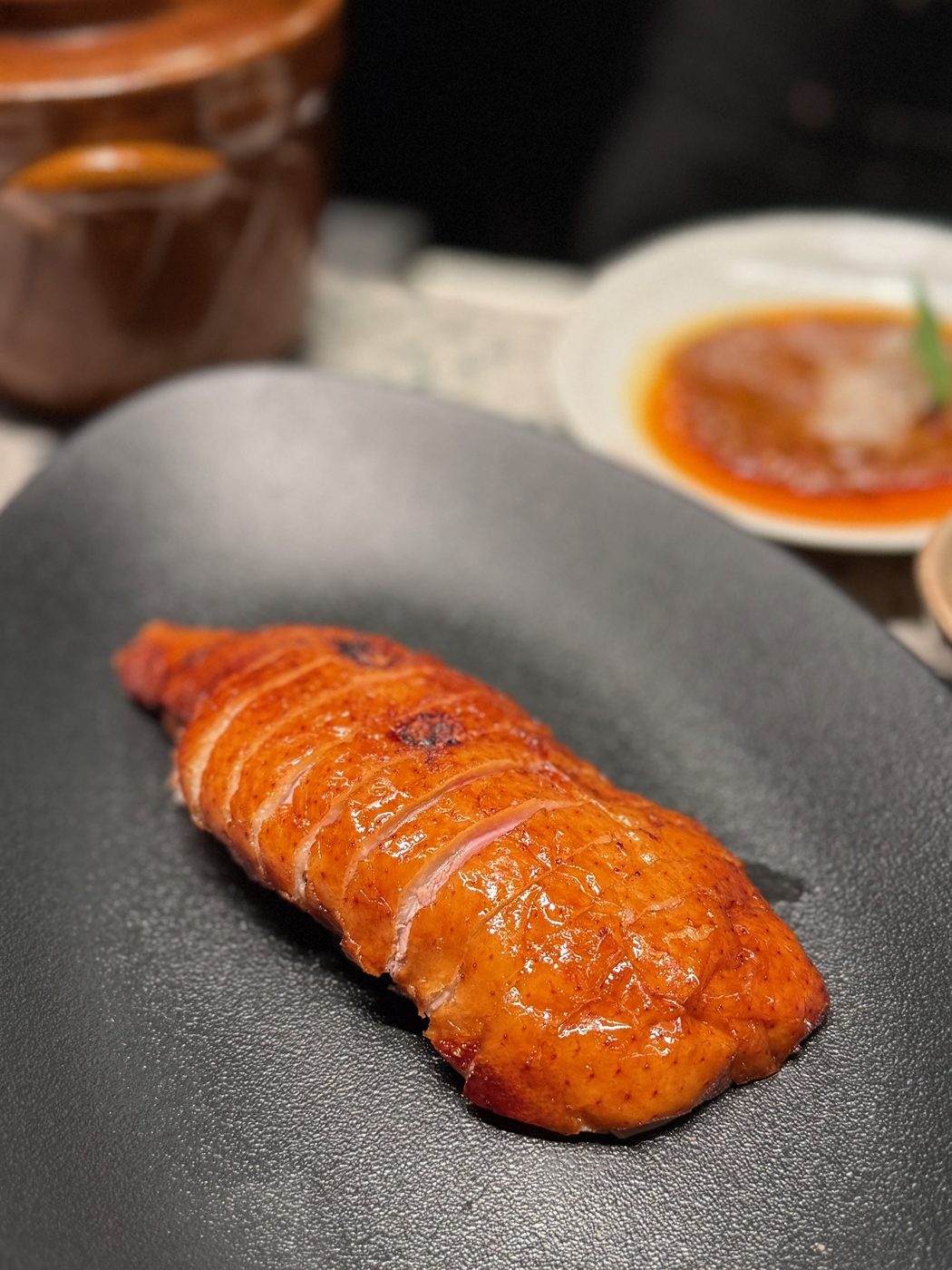 Sliced roasted duck breast with lacquered, scored skin on a matte black plate, a clay pot and a dish of sauce behind