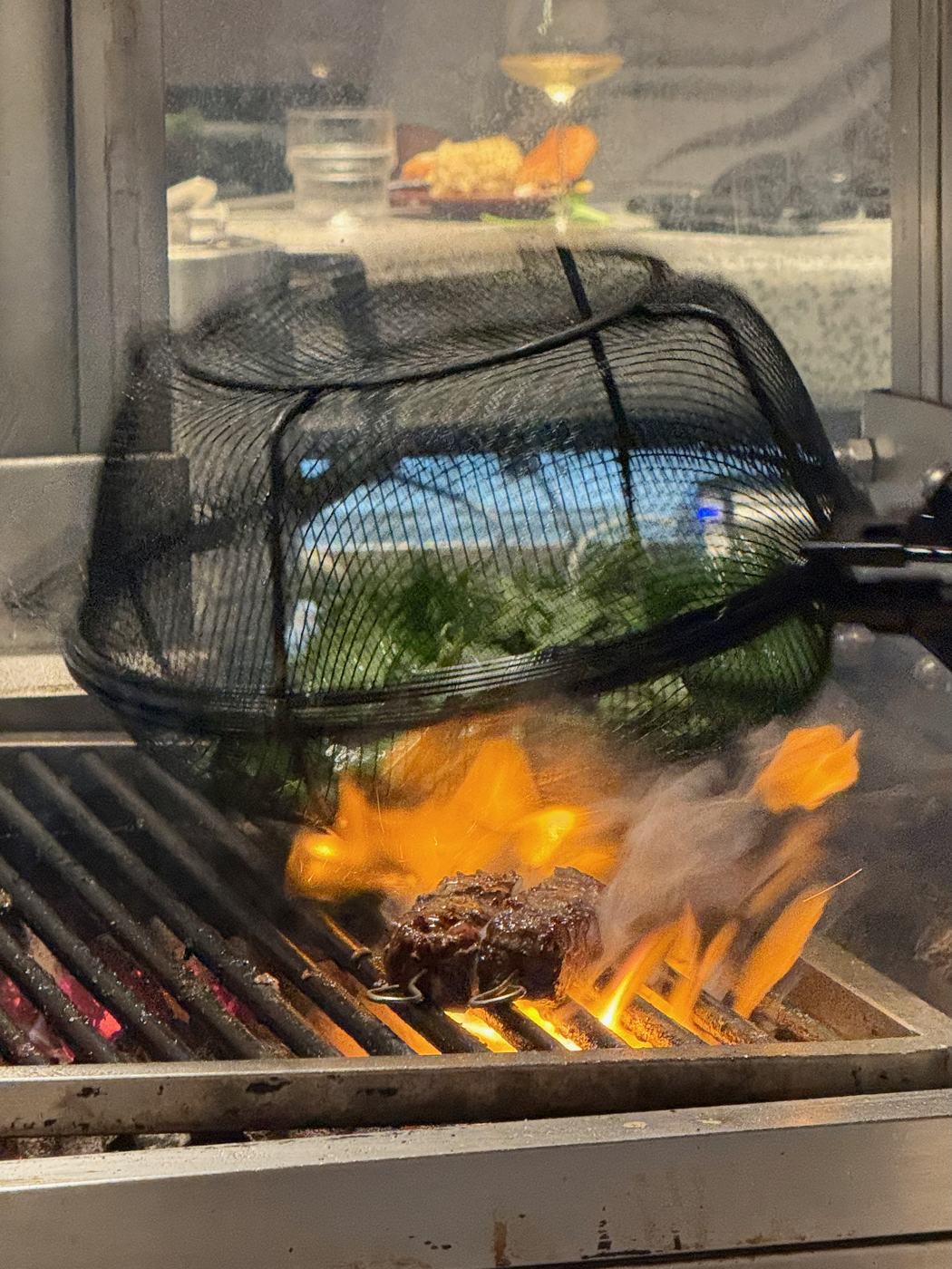 A wire basket of greens held over a charcoal grill, flames rising around skewered beef below, the dining room reflected in the glass behind