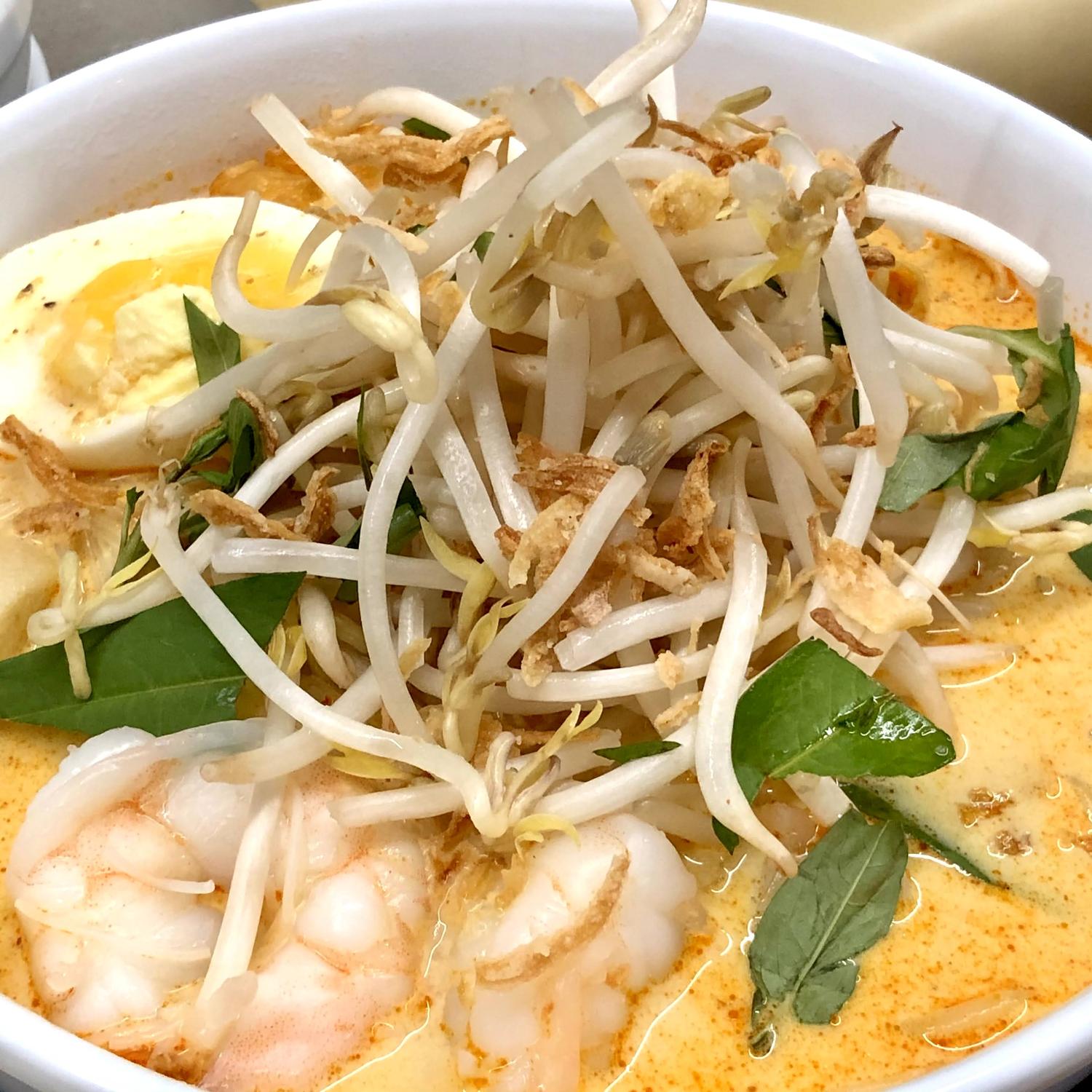 A bowl of laksa in an orange coconut broth, topped with prawns, a halved boiled egg, tofu puff, a heap of bean sprouts, laksa leaf and crisp fried shallot