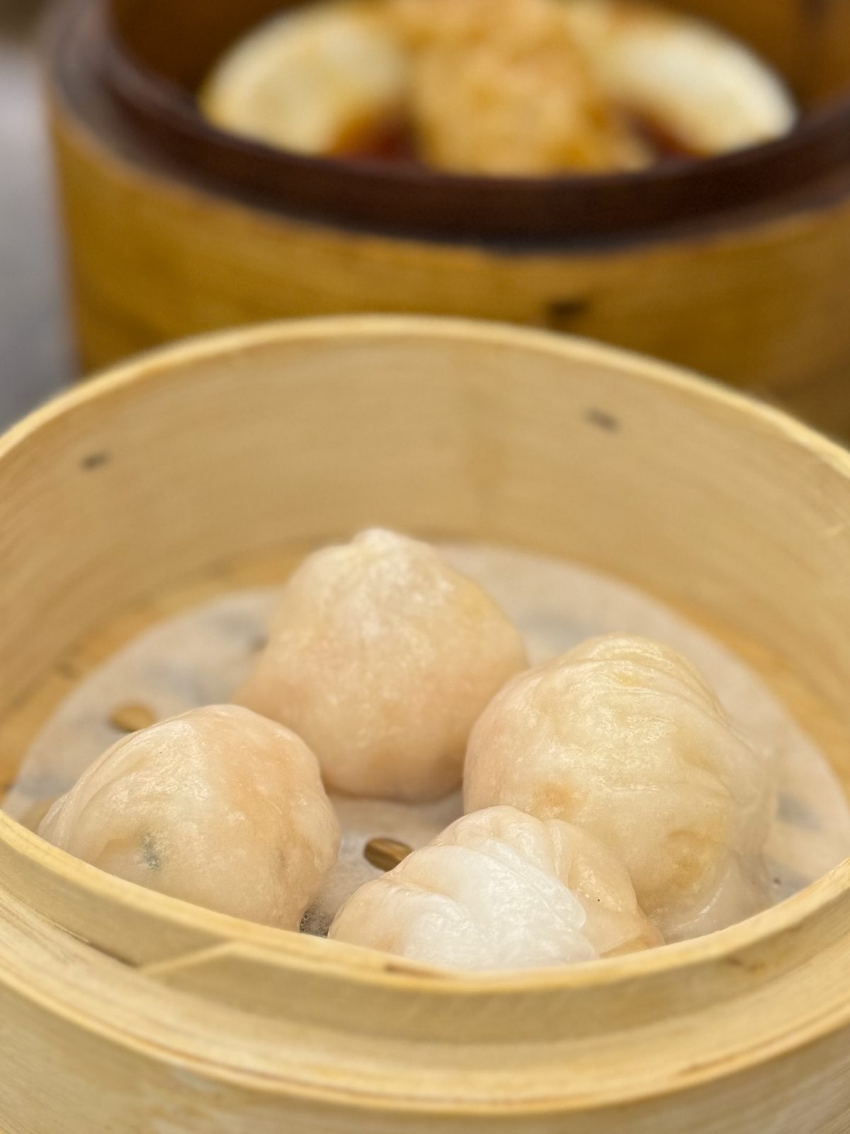 Four har gow in a bamboo steamer, their pleated translucent wrappers showing the pink prawn inside, a second steamer of buns behind