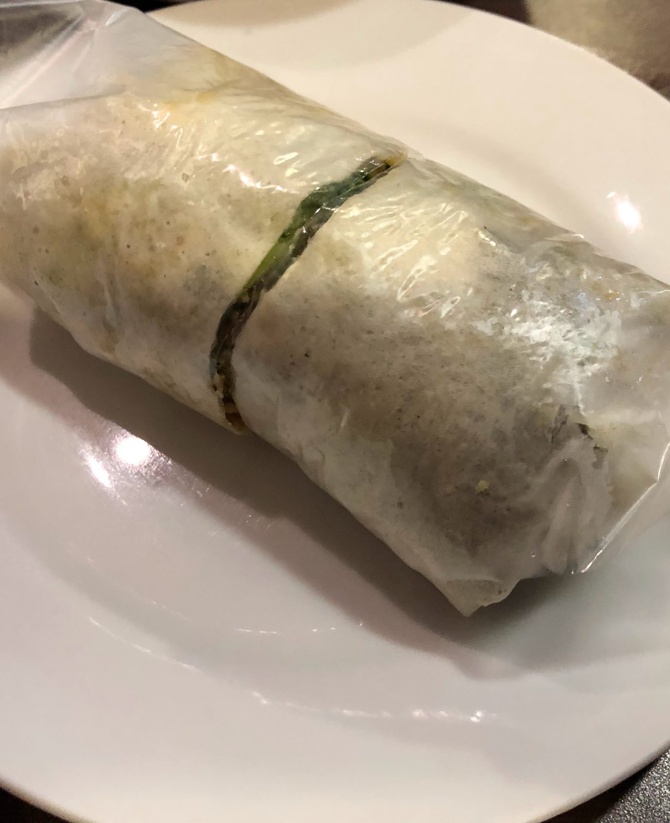 A single fresh lumpia on a white plate, its soft pale wrapper rolled around a filling with a strip of green leaf showing at the seam