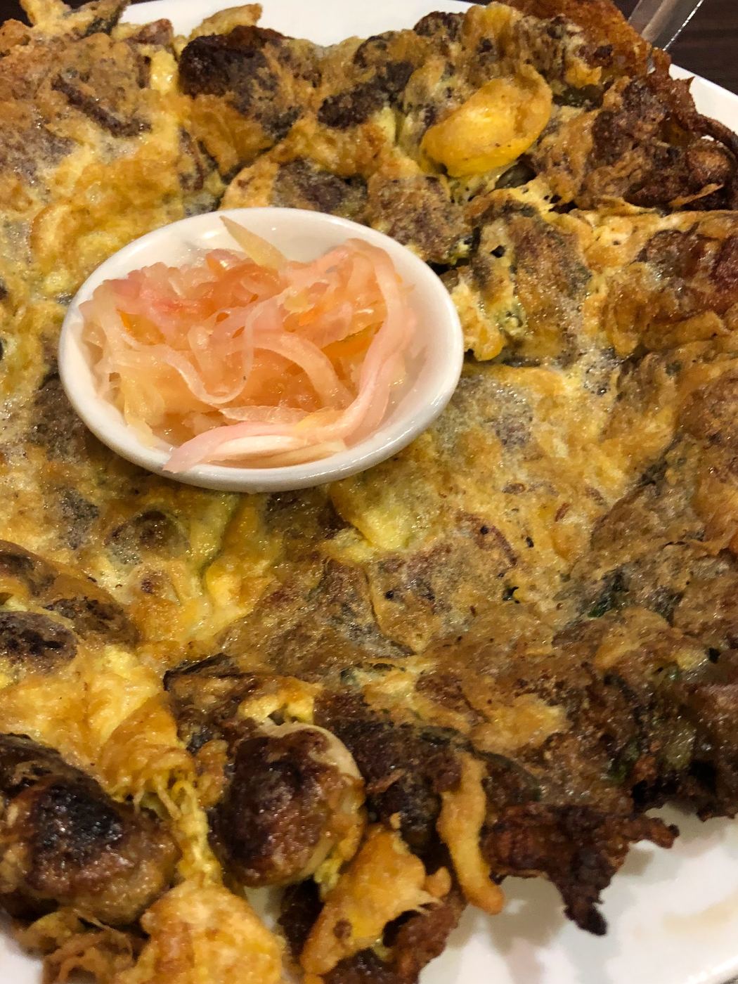 A large oyster omelette filling the plate, egg set around oysters and crisped brown at the edges, with a small dish of pickled onion and tomato at the centre