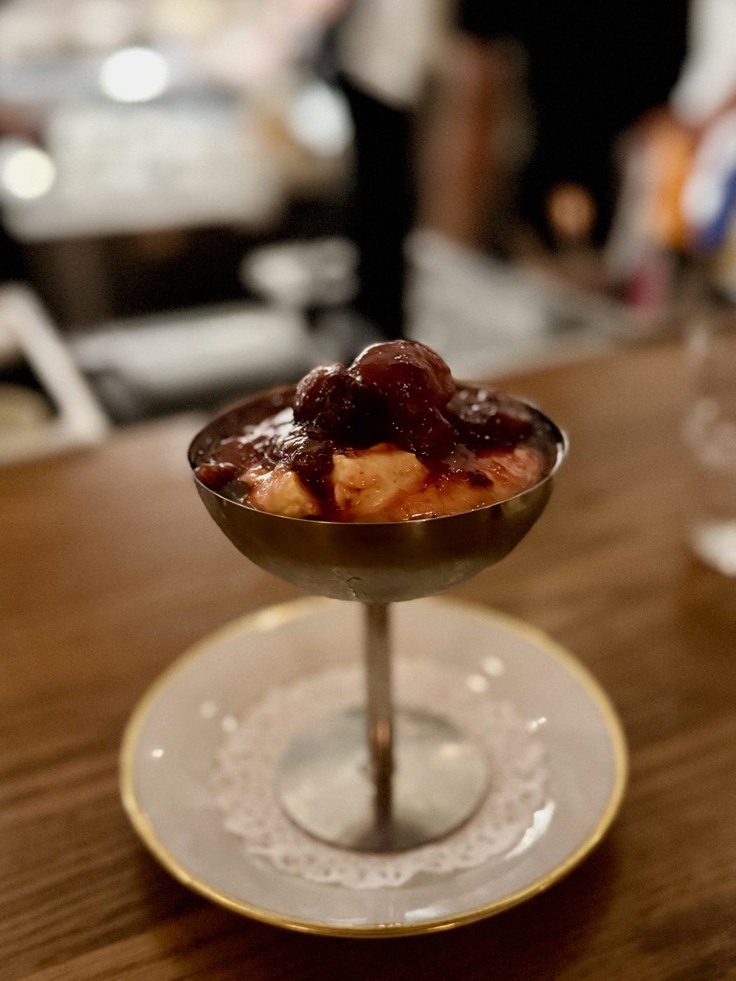 A footed metal coupe on a gold-rimmed saucer holding pale cream under a heap of dark red poached plums
