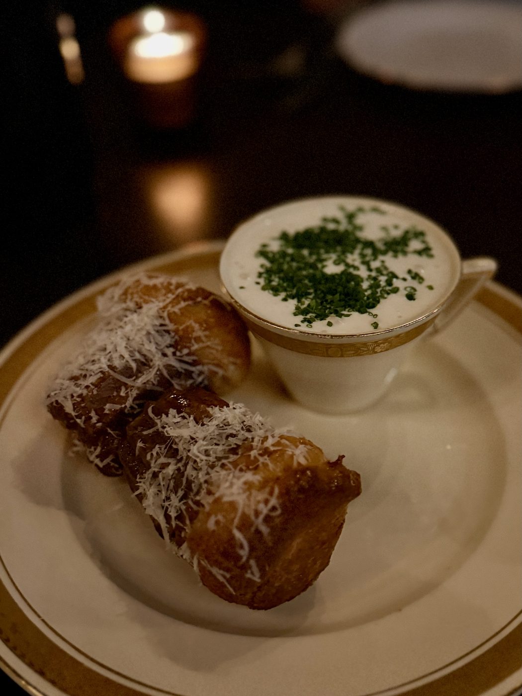 Two golden puffs blanketed in finely grated parmesan on a gold-rimmed plate, with warm parmesan mousse scattered with chopped chives in a gold-rimmed teacup alongside